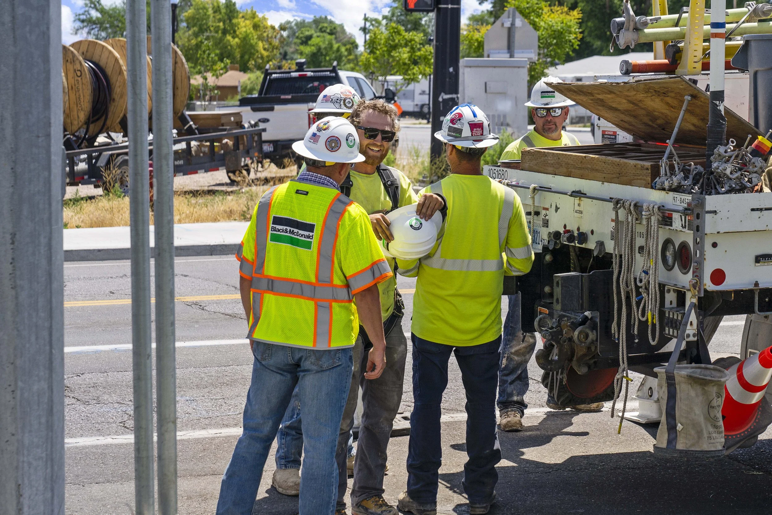 Group of construction workers in bright yellow safety vests and white hard hats gathered around a utility truck on a street with trees in the background.