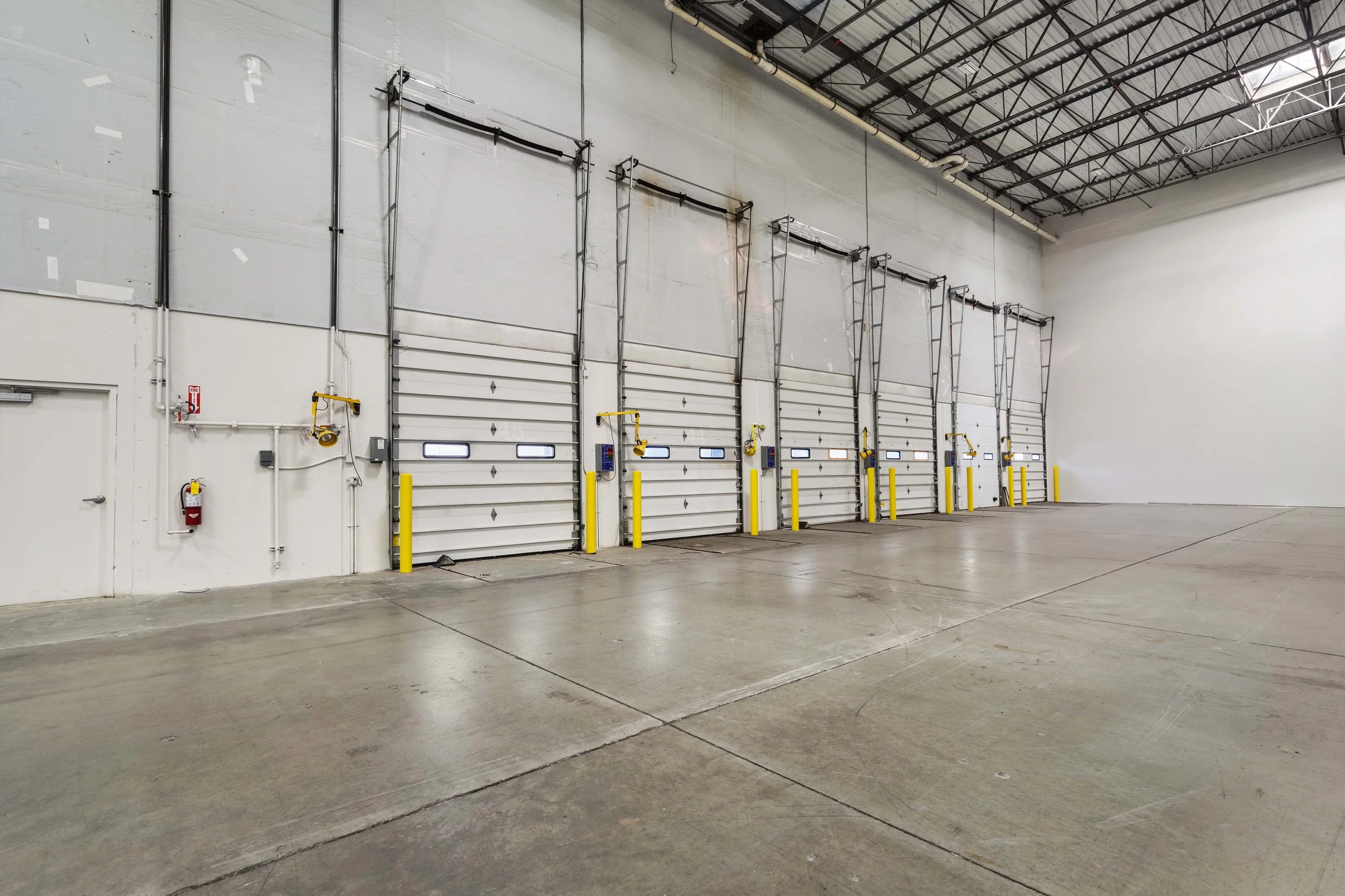 An empty warehouse loading dock with five roll-up garage doors, each equipped with yellow safety bollards, automatic openers, and small windows at the top.