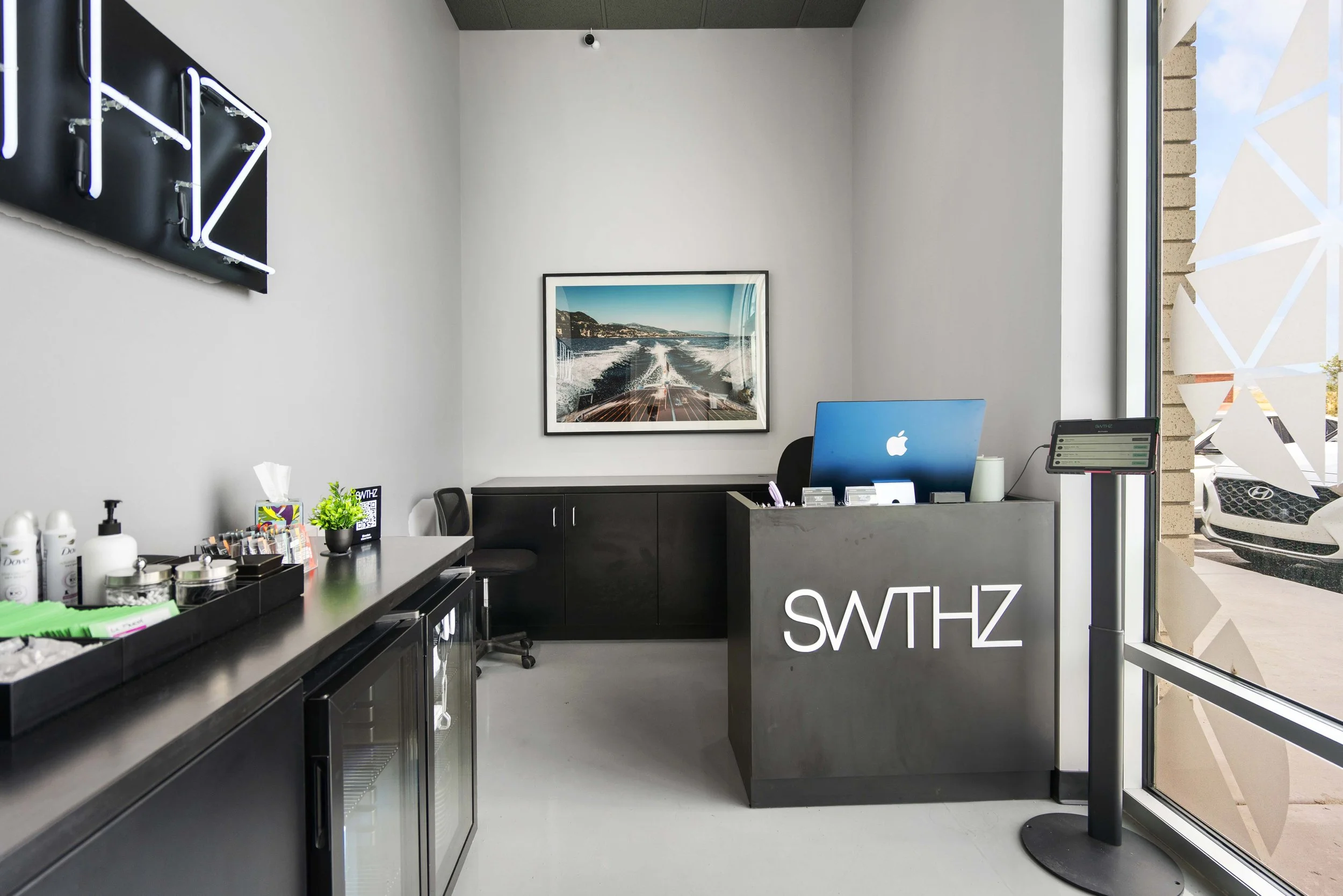 Modern reception area with black counter displaying the logo 'SWITHZ', a computer, and a tablet, along with a black cabinet, chairs, wall art of a boat on water, sanitizer bottles, tissues, and toiletries, with a large window showing parked cars outs