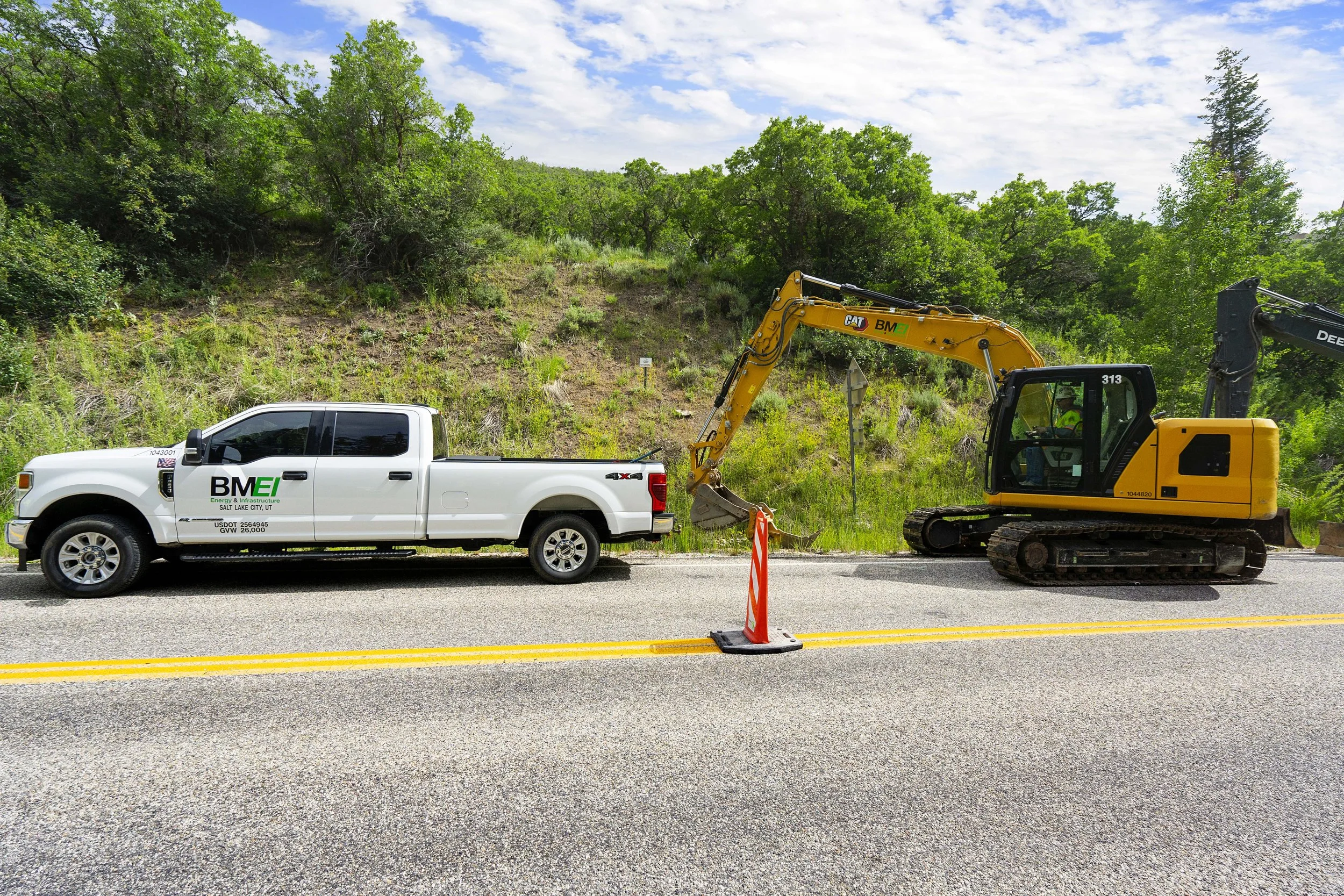 A white truck with BME logo connected to a yellow excavator on a road, with construction barricade and a hillside with trees in the background.