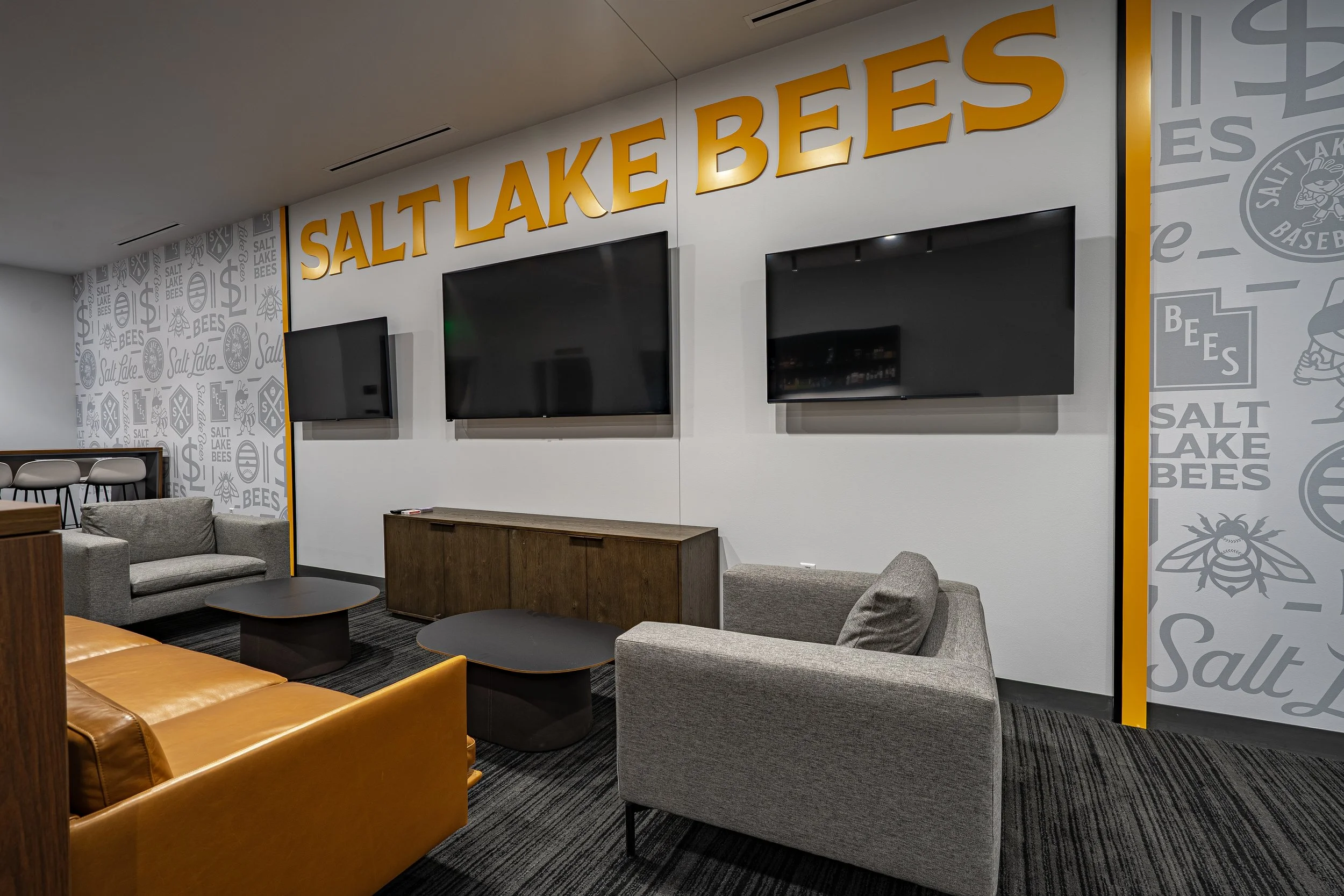 A lounge area with gray and orange furniture, including two sofas and two coffee tables, against a white wall with three mounted TVs and signage reading 'Salt Lake Bees'.