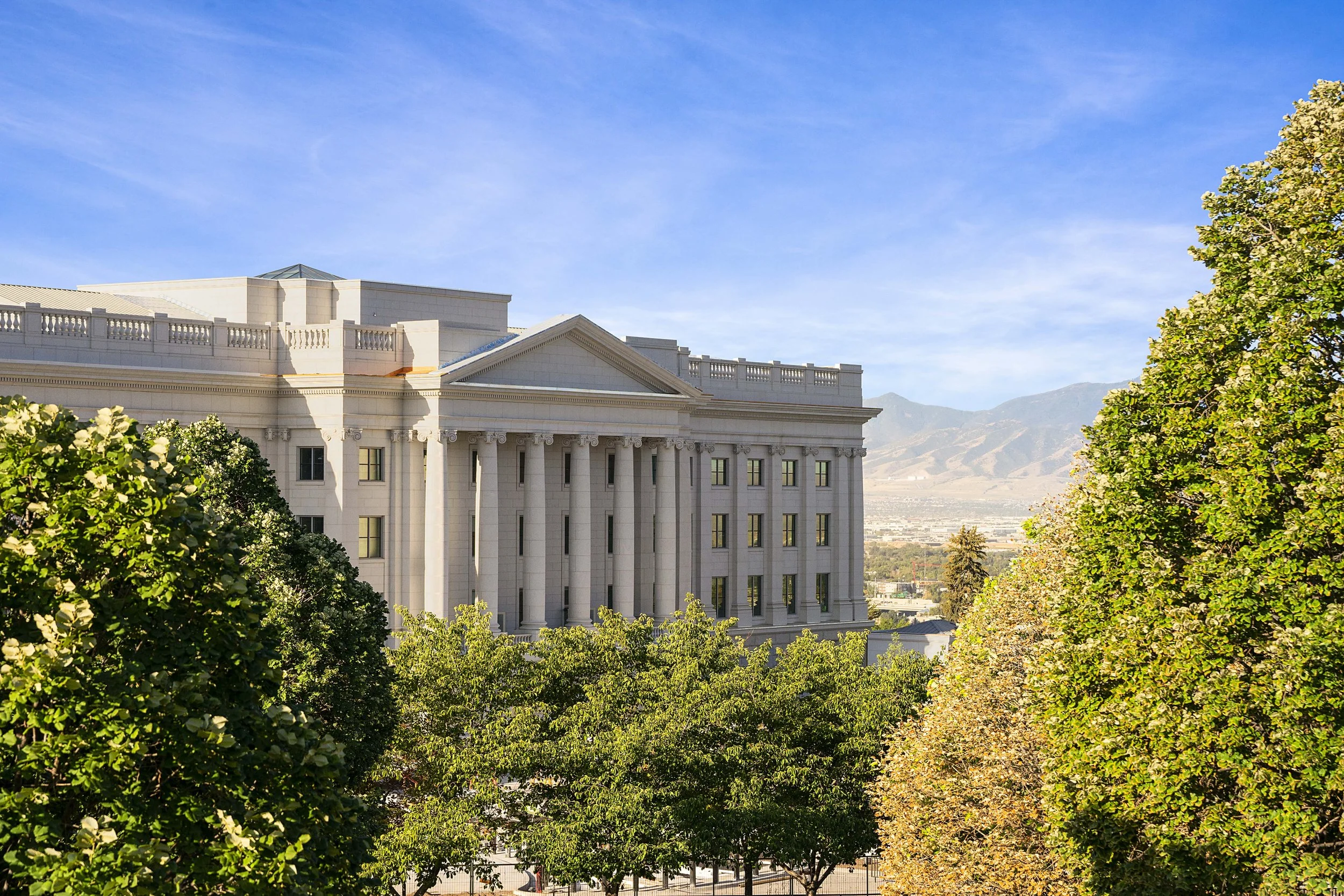View of a large white government or institutional building with columns, surrounded by green trees, with mountains and a cityscape in the background under a clear blue sky.