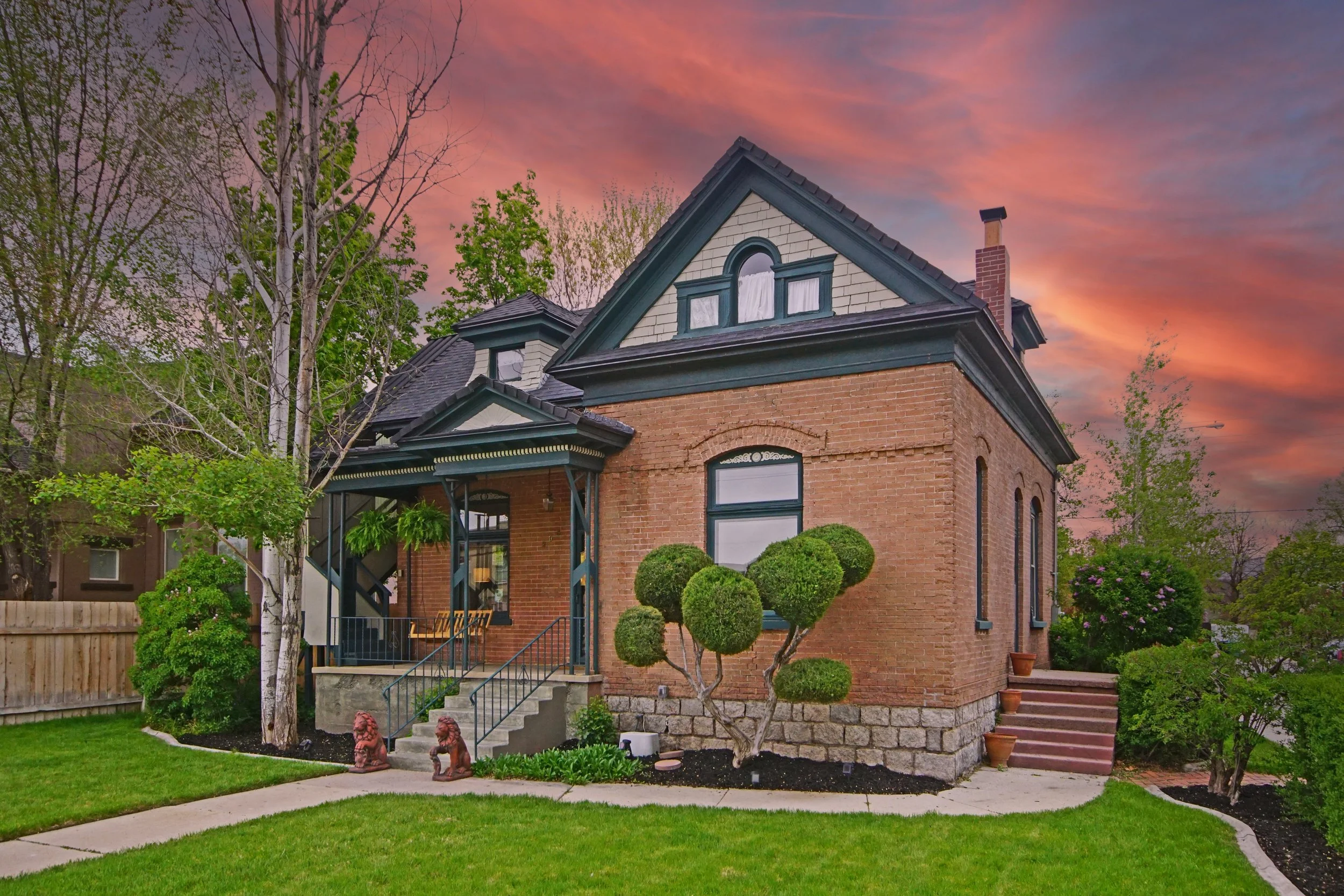 Victorian-style brick house with landscaped front yard, manicured shrubs, and a sunset sky.
