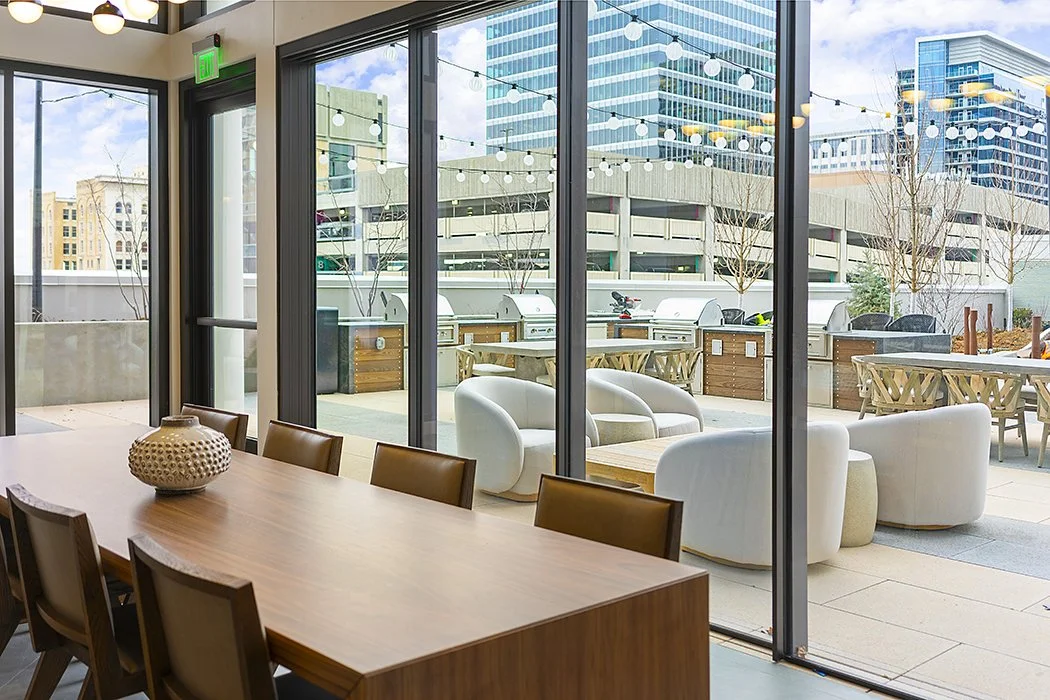 Indoor dining area with large glass windows opening to an outdoor patio with seating, string lights, and cityscape in the background.