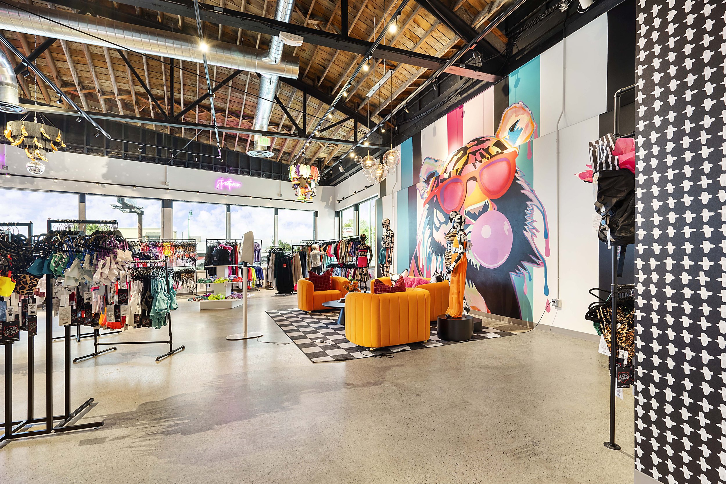 Interior of a colorful clothing store with clothing racks, mannequins, orange sofas, and a vibrant wall mural of a bear wearing sunglasses.