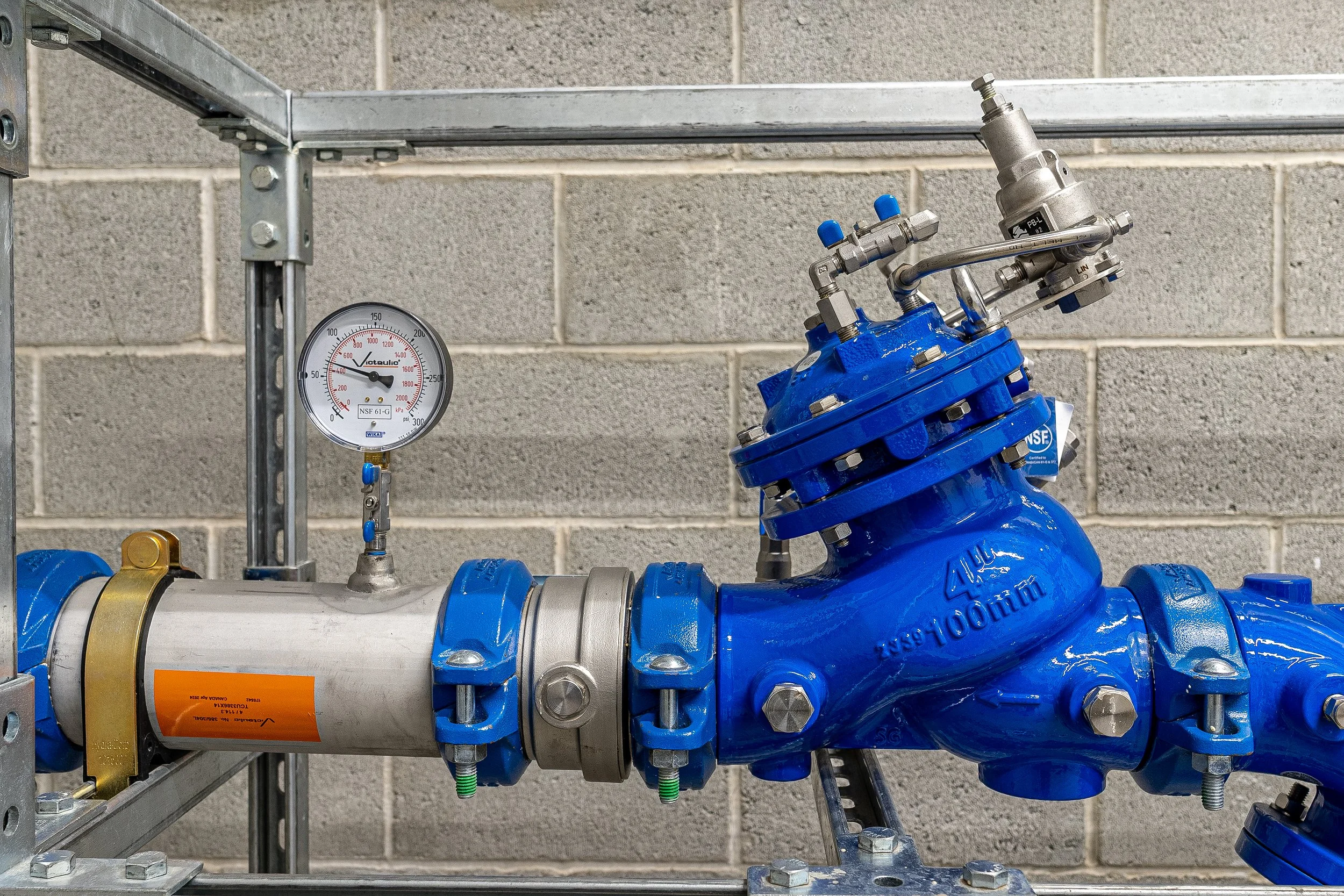 Industrial pipeline system with a pressure gauge and blue valve assembly against a concrete block wall.