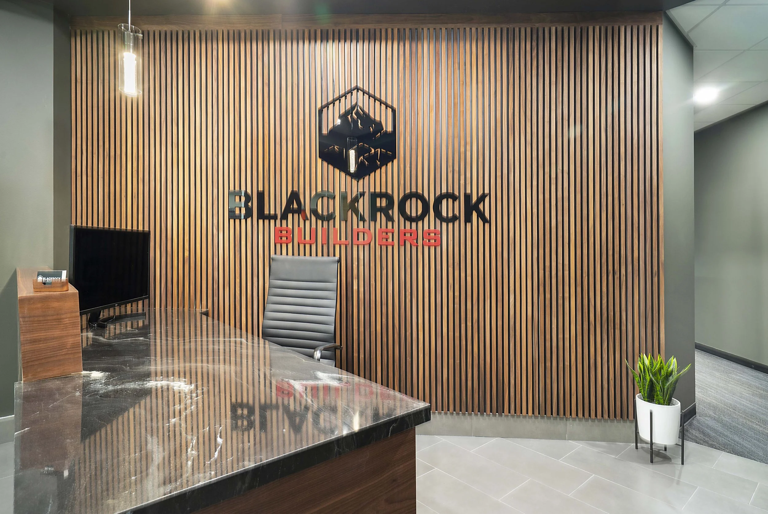 Office reception area with a wooden slat wall, sign reading "BLACKROCK BUILDERS," a black office chair, a desk with a marble top, a computer monitor, a potted plant, and soft lighting.