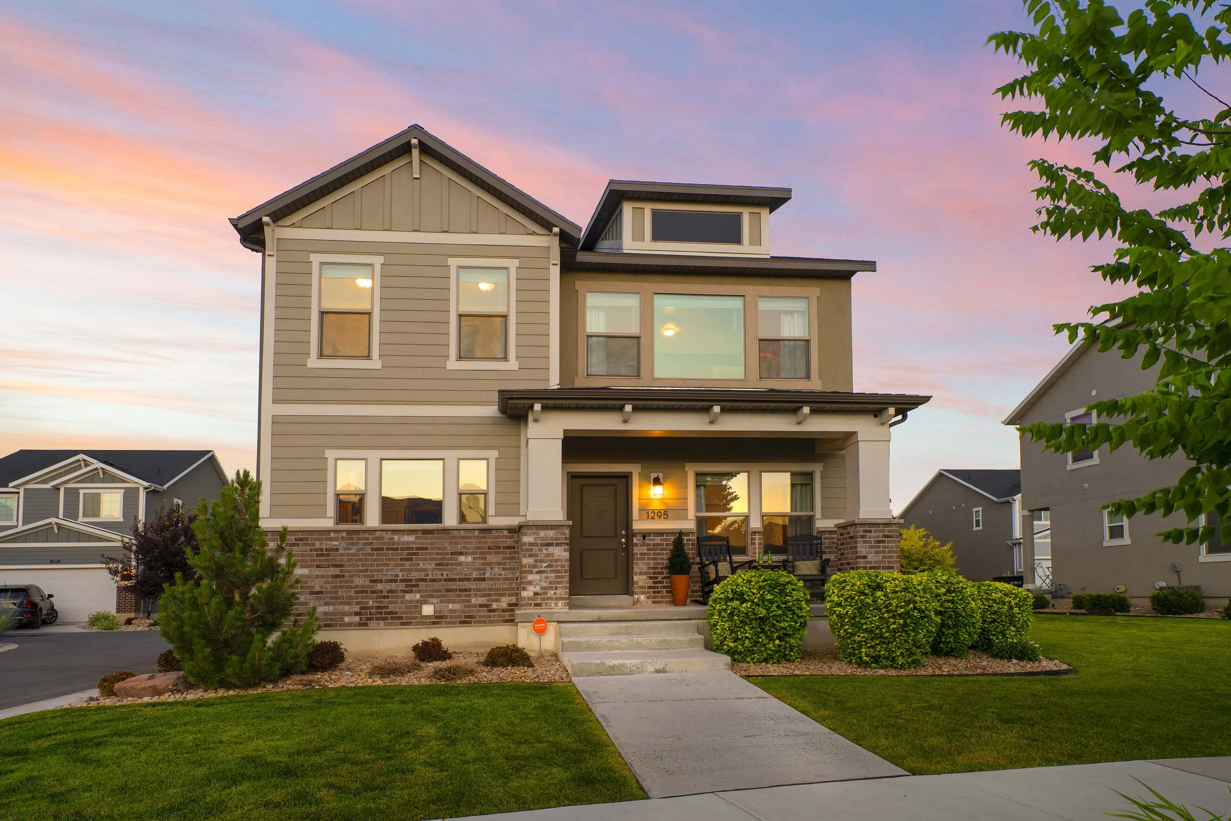 A modern, three-story house with a front porch, brick and siding exterior, surrounded by a neatly landscaped lawn with bushes and trees, during sunset.
