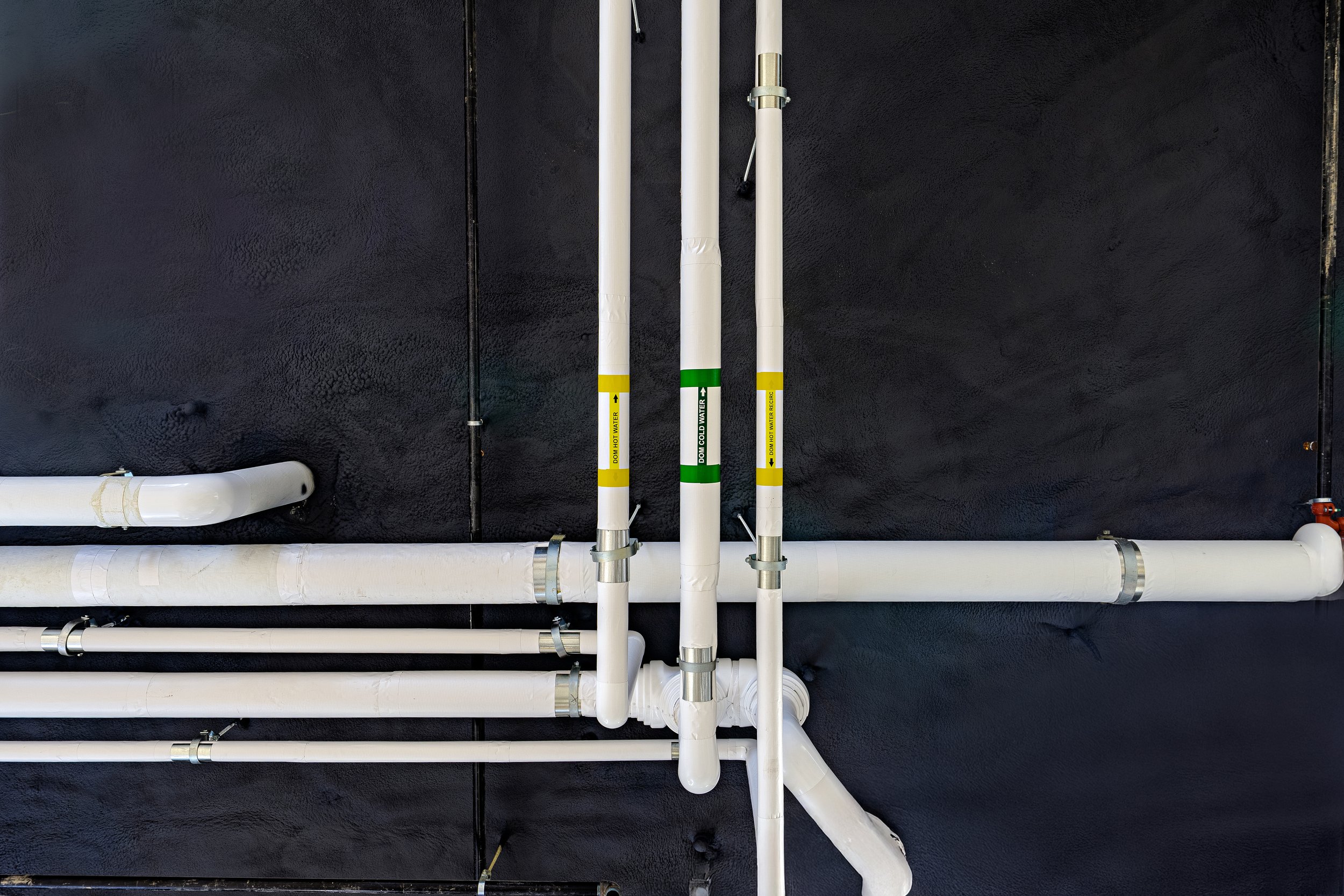 Multiple white pipes with labels and metal clamps are installed on a black textured wall, some pipes are joined at angles and have signs indicating water flow directions.
