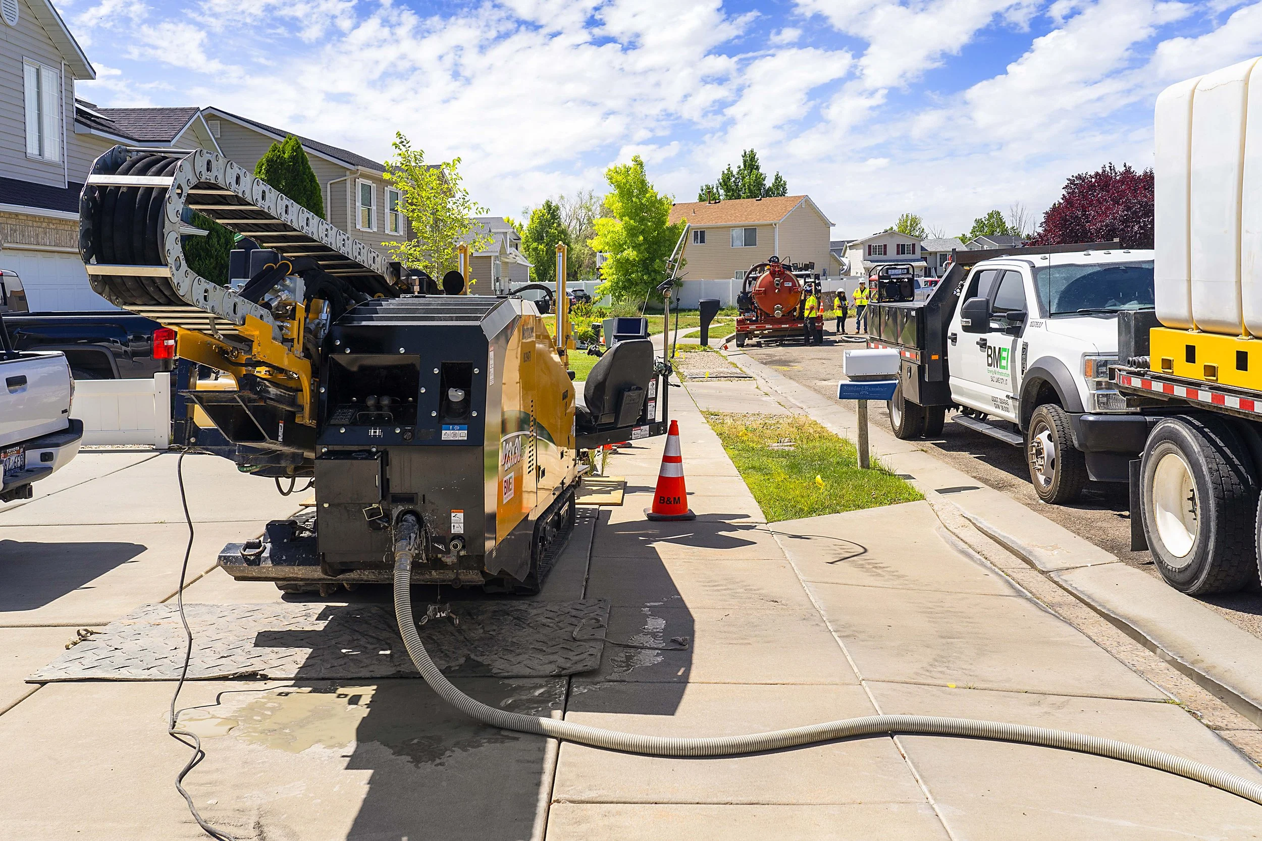 Construction work on a residential street with machinery, trucks, and workers, clear blue sky, and houses in the background.