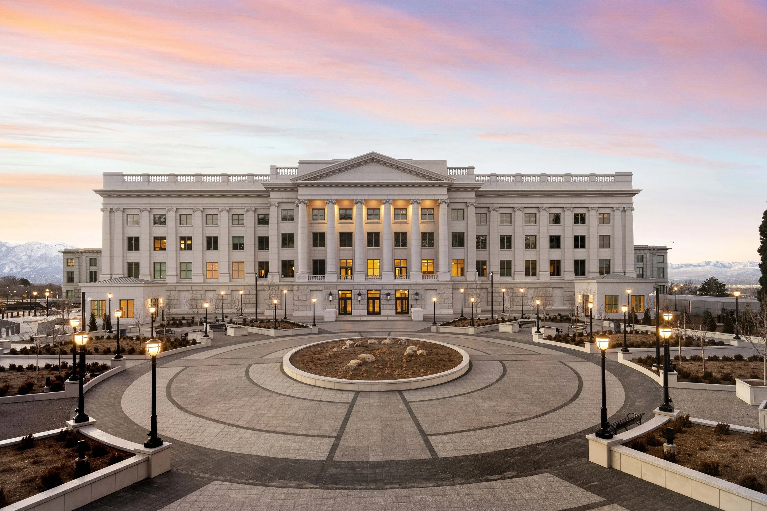 A large white government or institutional building with classical architecture, including columns and a pediment, is illuminated by streetlights at dusk. The building is surrounded by a paved courtyard with circular patterns and landscaped areas.