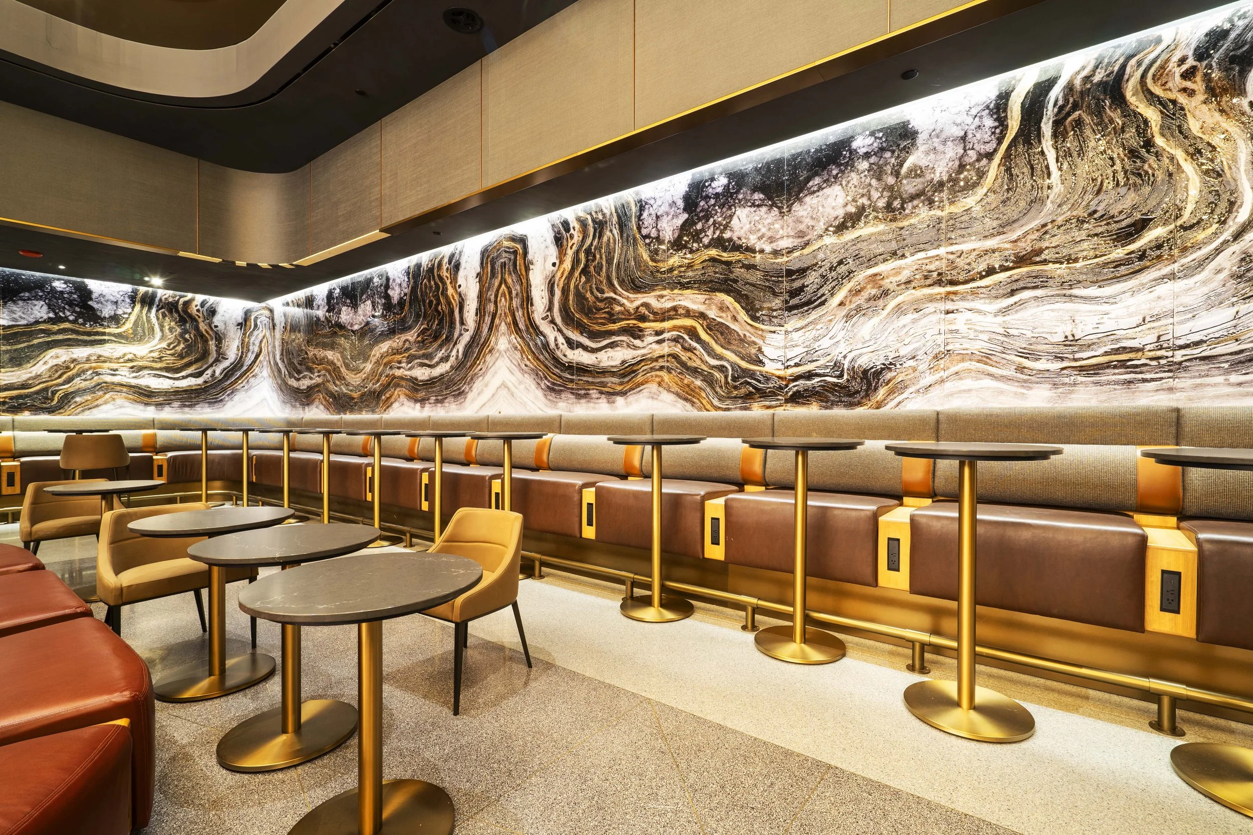 Interior of a modern lounge with a large marble-patterned wall, small round tables, and cushioned chairs with electrical outlets.