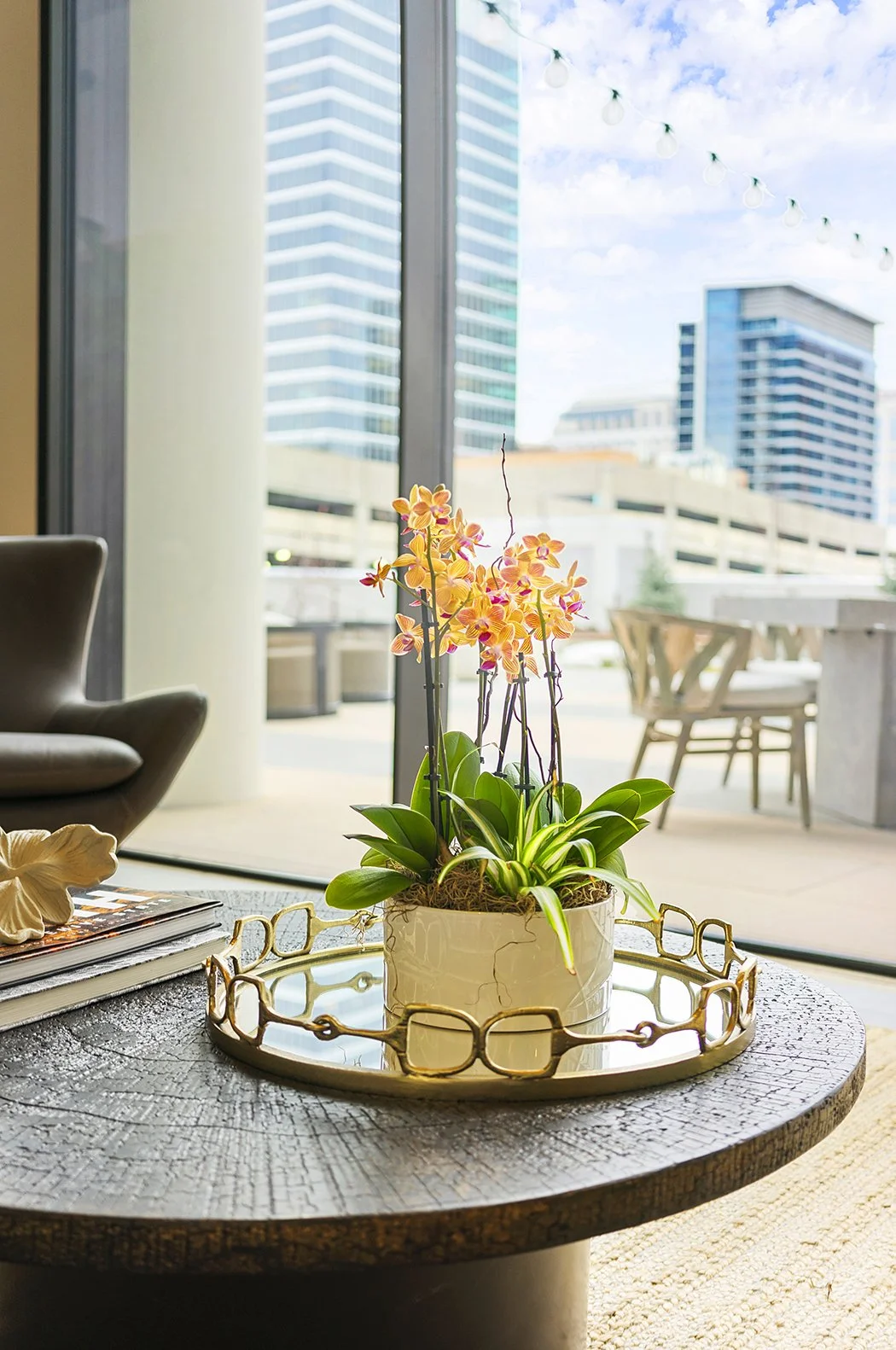 A potted orchid plant with pink and yellow flowers on a round wooden table inside a modern apartment, with a cityscape and large glass windows in the background.