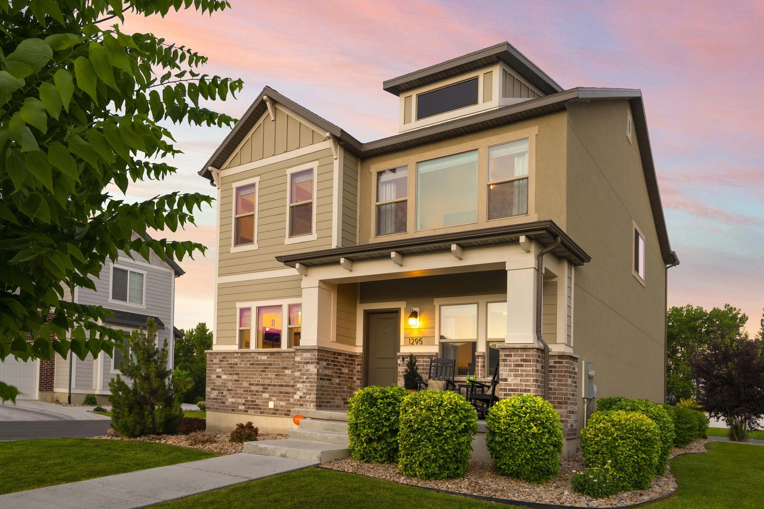 A modern, two-story house with a mix of beige and gray exterior, brick accents, large windows, and a small front porch with chairs, surrounded by green shrubs and trees, during sunset.