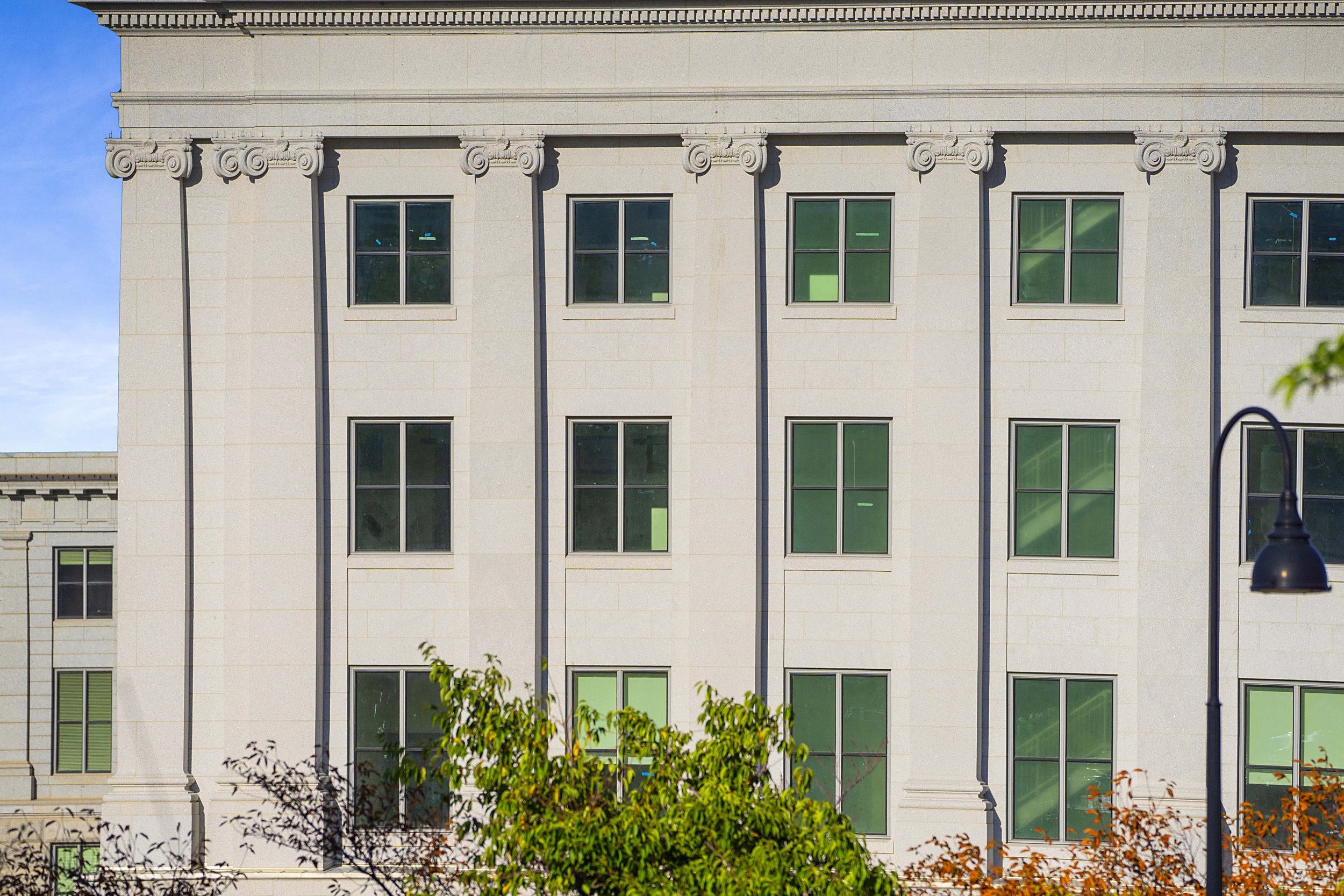 A multi-story beige stone building with symmetrical windows and decorative architectural details, partly obscured by green trees and a street lamp in the foreground.