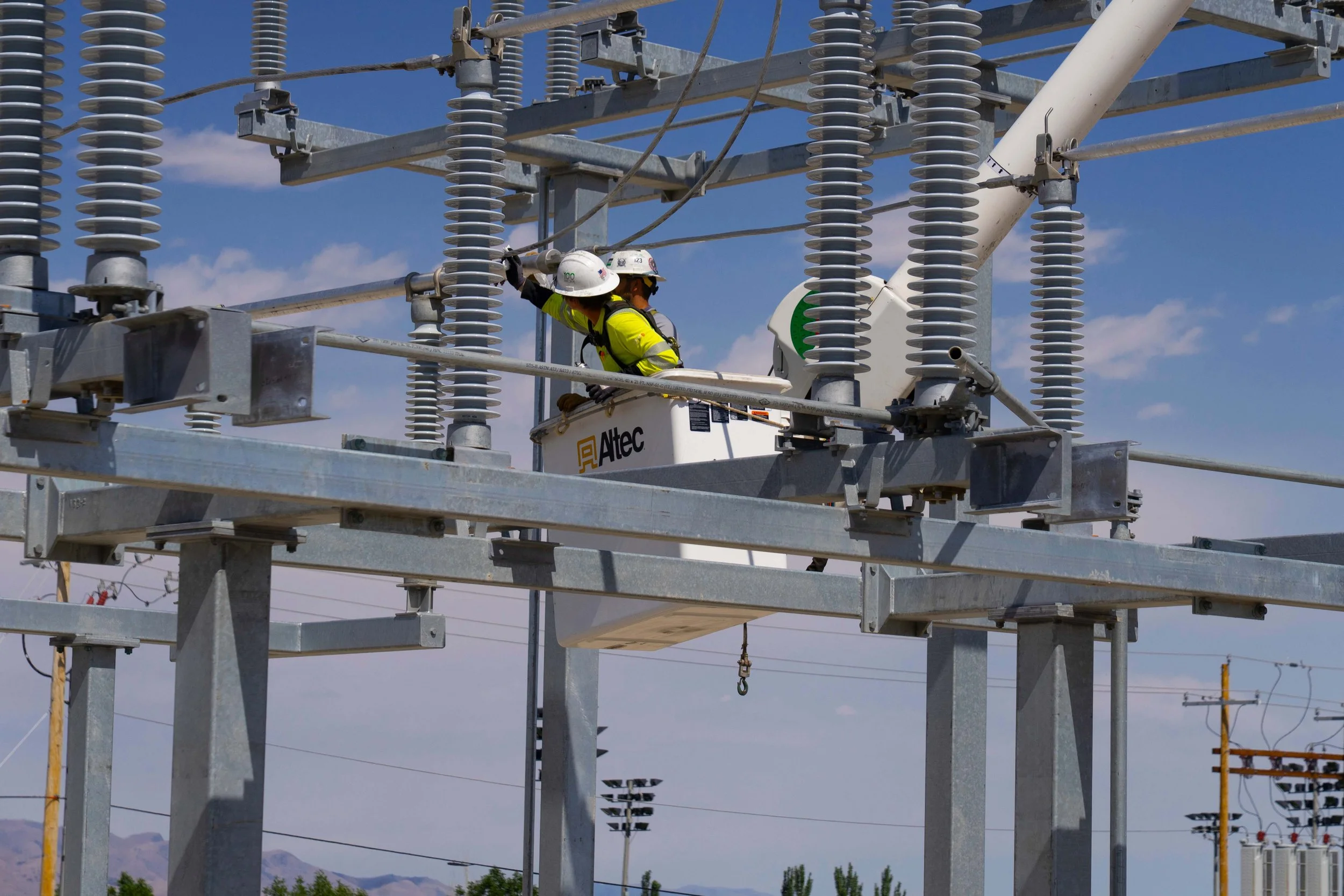Two workers in safety gear working on electrical equipment at an outdoor power station or substation, surrounded by high-voltage insulators and steel structures.