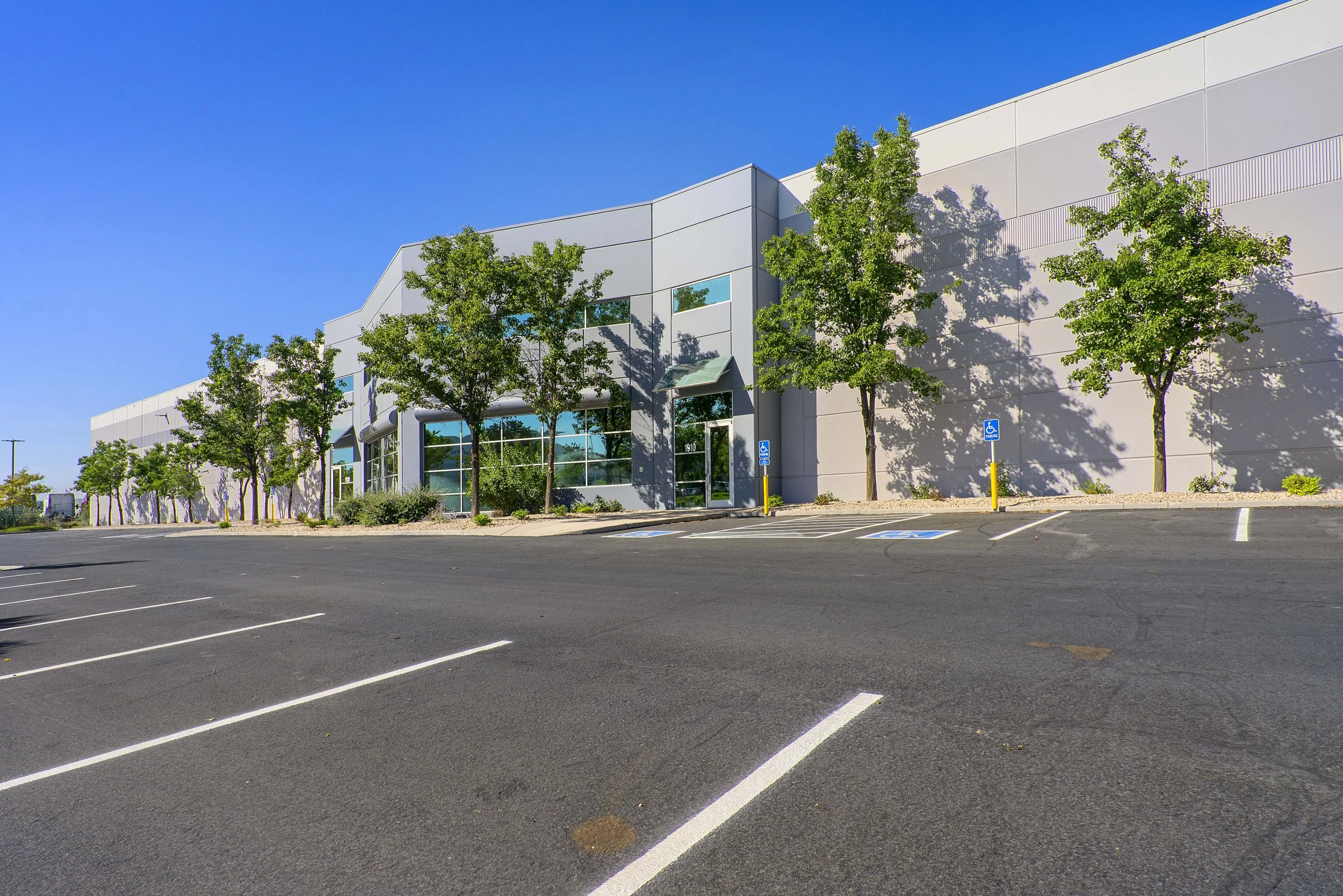 Empty parking lot in front of a modern commercial building with several trees and clear blue sky.