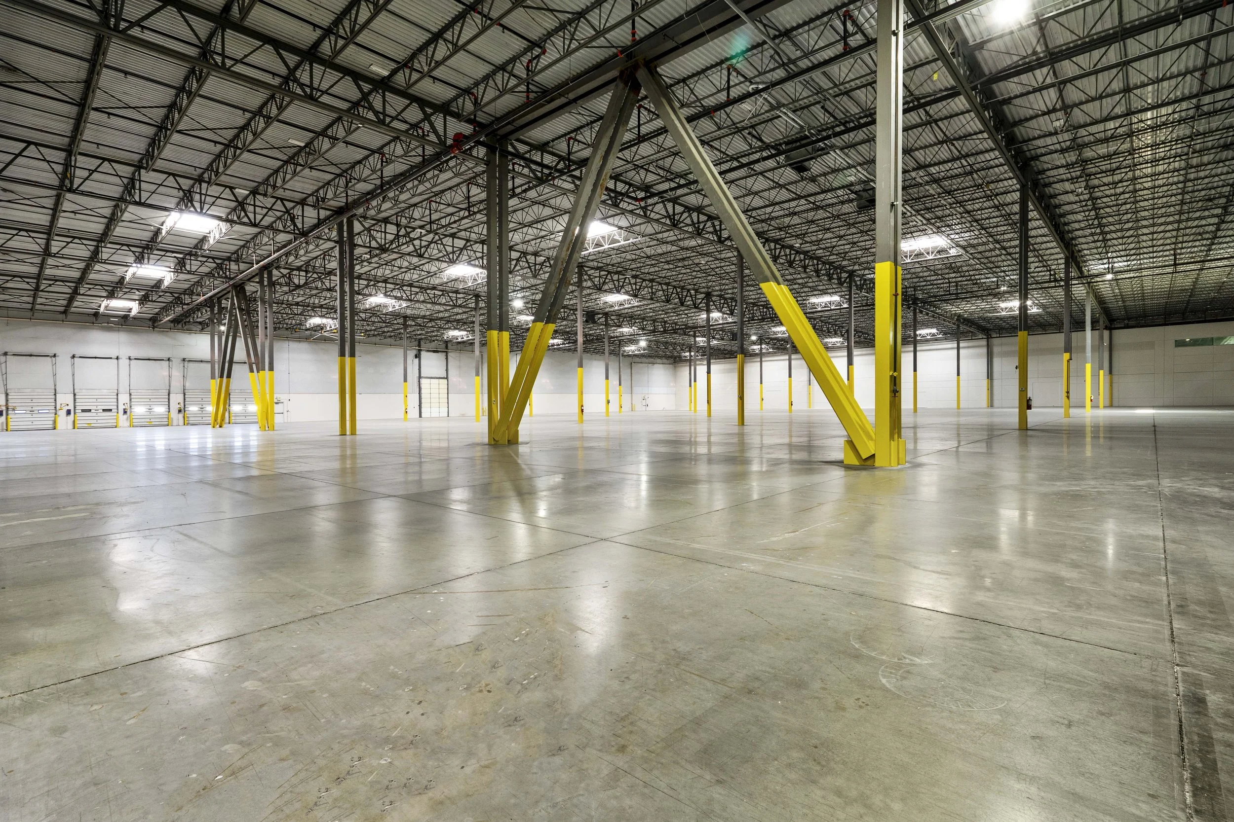 Empty warehouse with concrete floors, metal ceiling, yellow safety posts, and overhead lighting.
