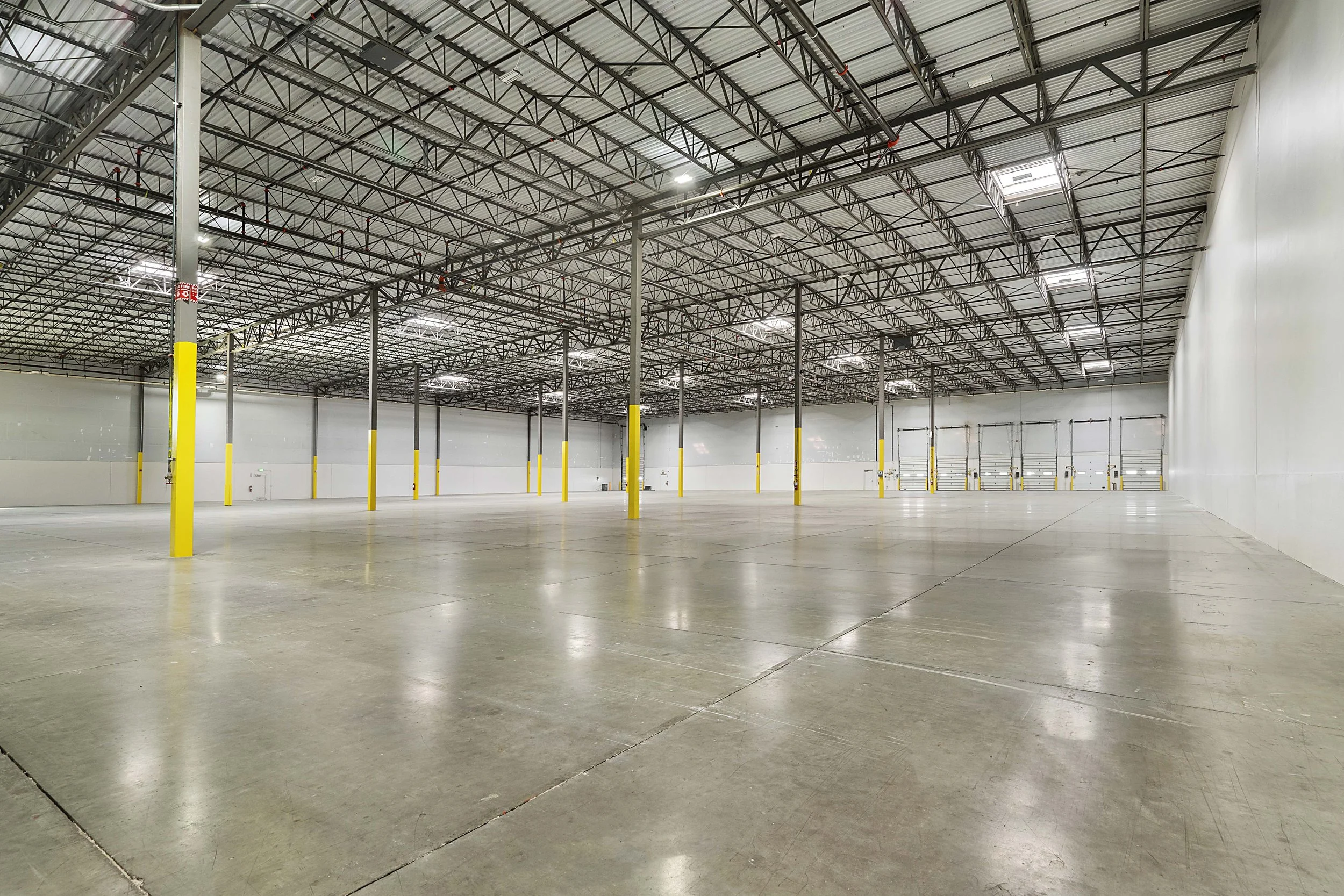 Empty warehouse with high ceiling, metal support beams, yellow safety posts, polished concrete floor, and loading dock doors in the distance.