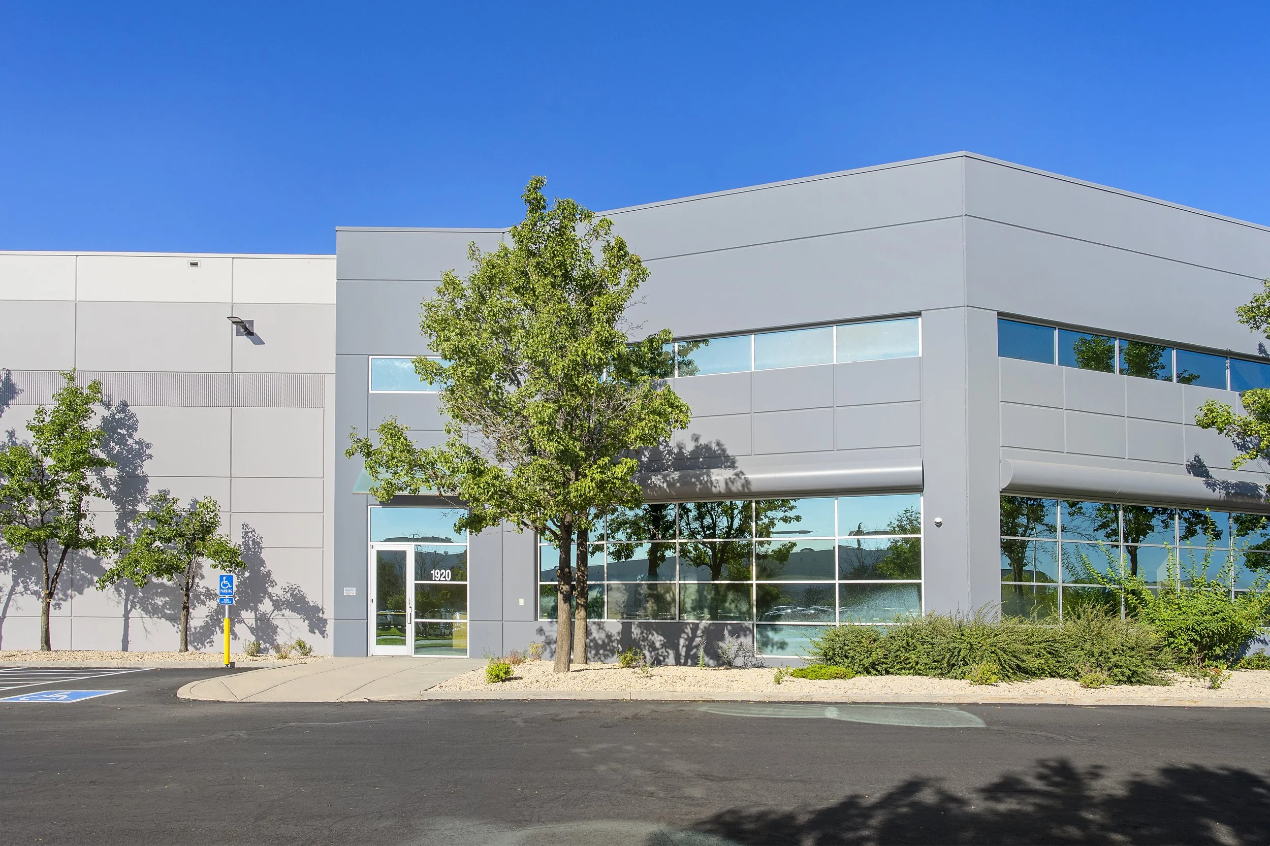 Modern grey commercial building with large glass windows, surrounded by trees and bushes, under a bright blue sky.