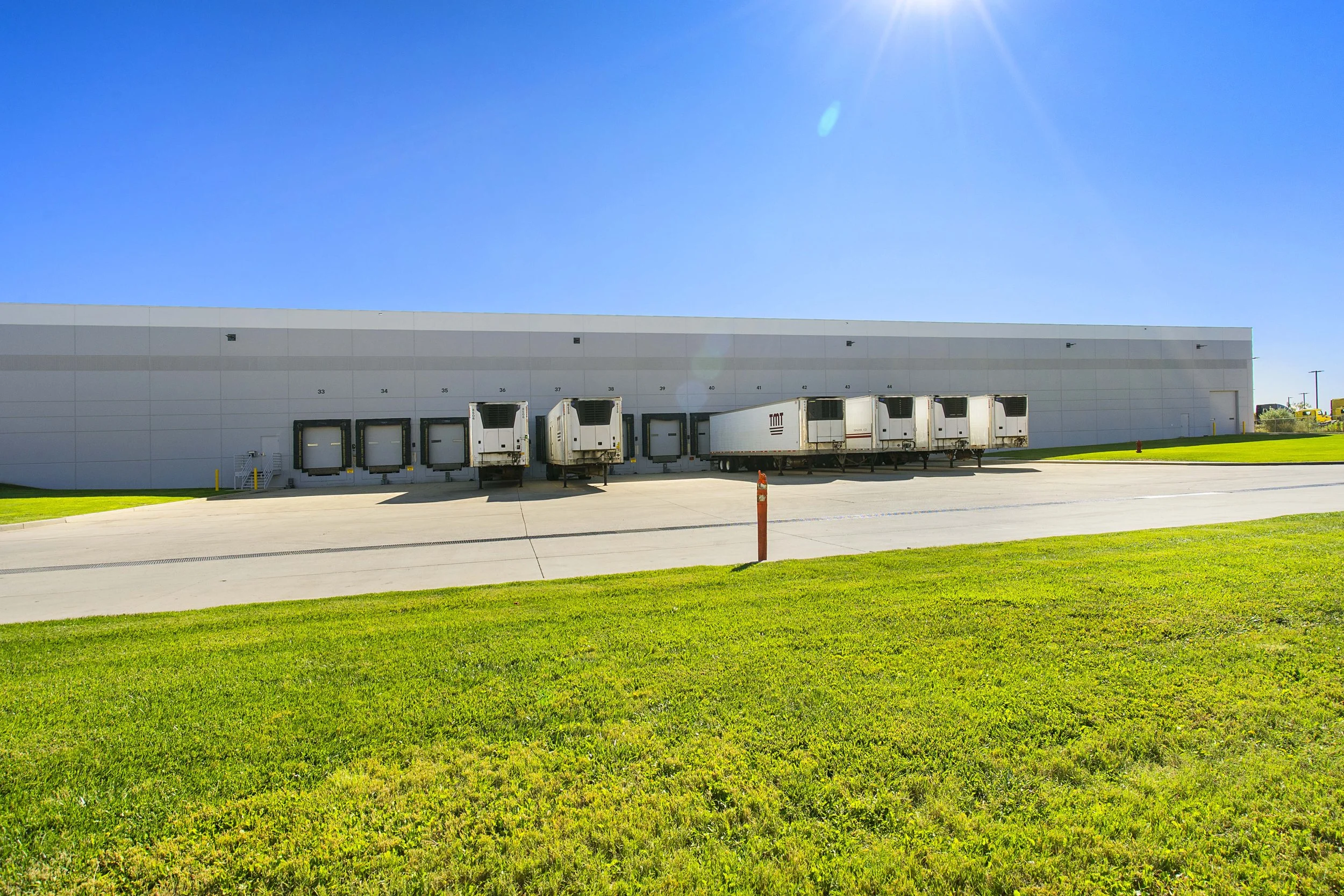 Large industrial warehouse with loading docks and trailers parked outside, surrounded by green grass and a bright blue sky.