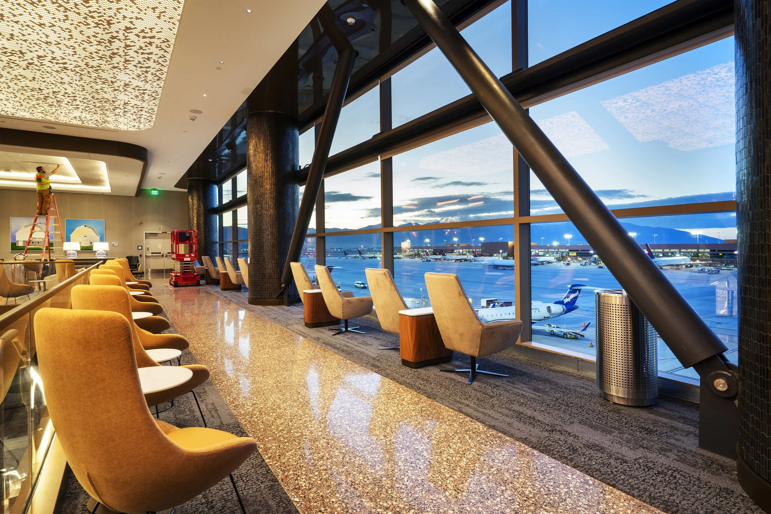 Airport terminal with large windows showing airplanes on the tarmac at sunset, with yellow and beige chairs along the window and a worker on a ladder installing or repairing ceiling lights.