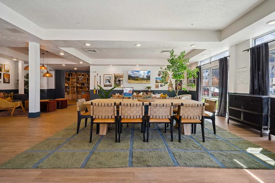 A spacious, brightly lit living or dining area with a large wooden table, eight chairs, an area rug, houseplants, wall art, and floor-to-ceiling windows with dark curtains.