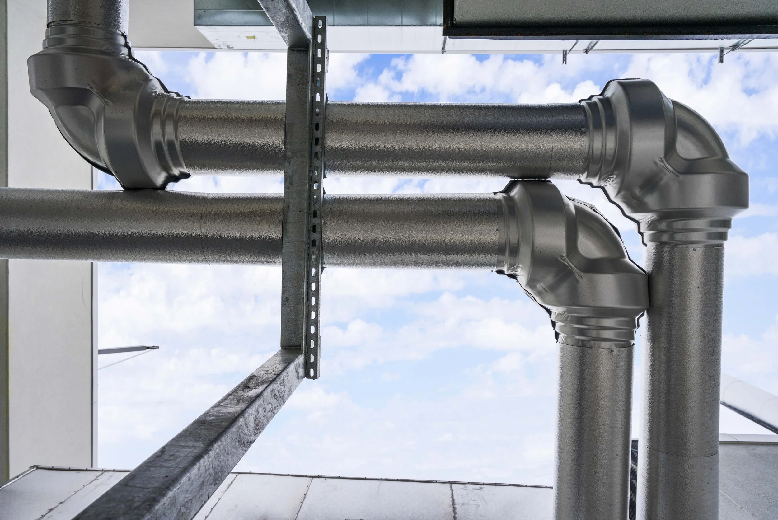 Metal industrial pipes with joints, installed outdoors against a sky with clouds.