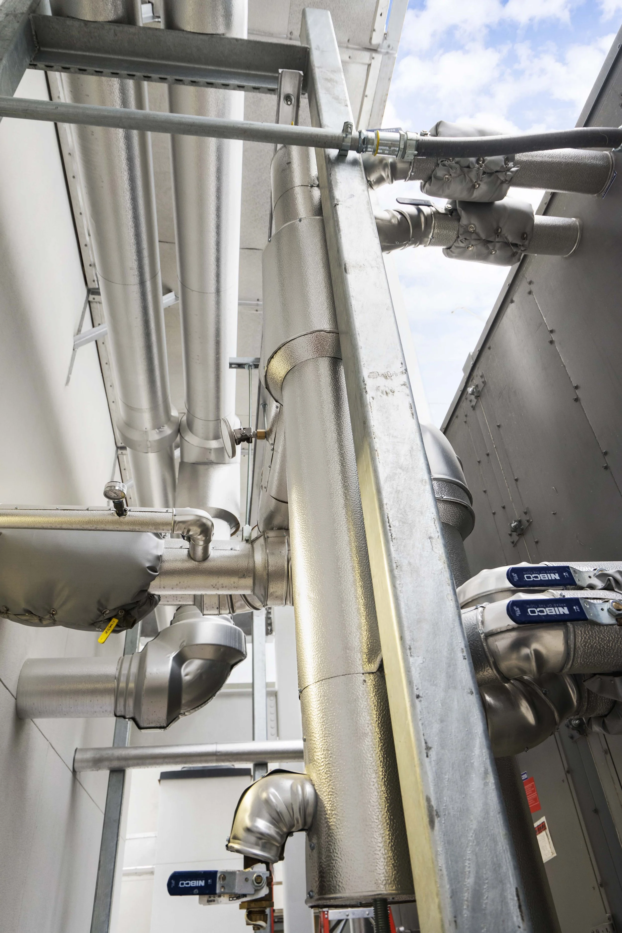 Industrial piping system with insulated pipes, valves, and metal support structure against a sky background.