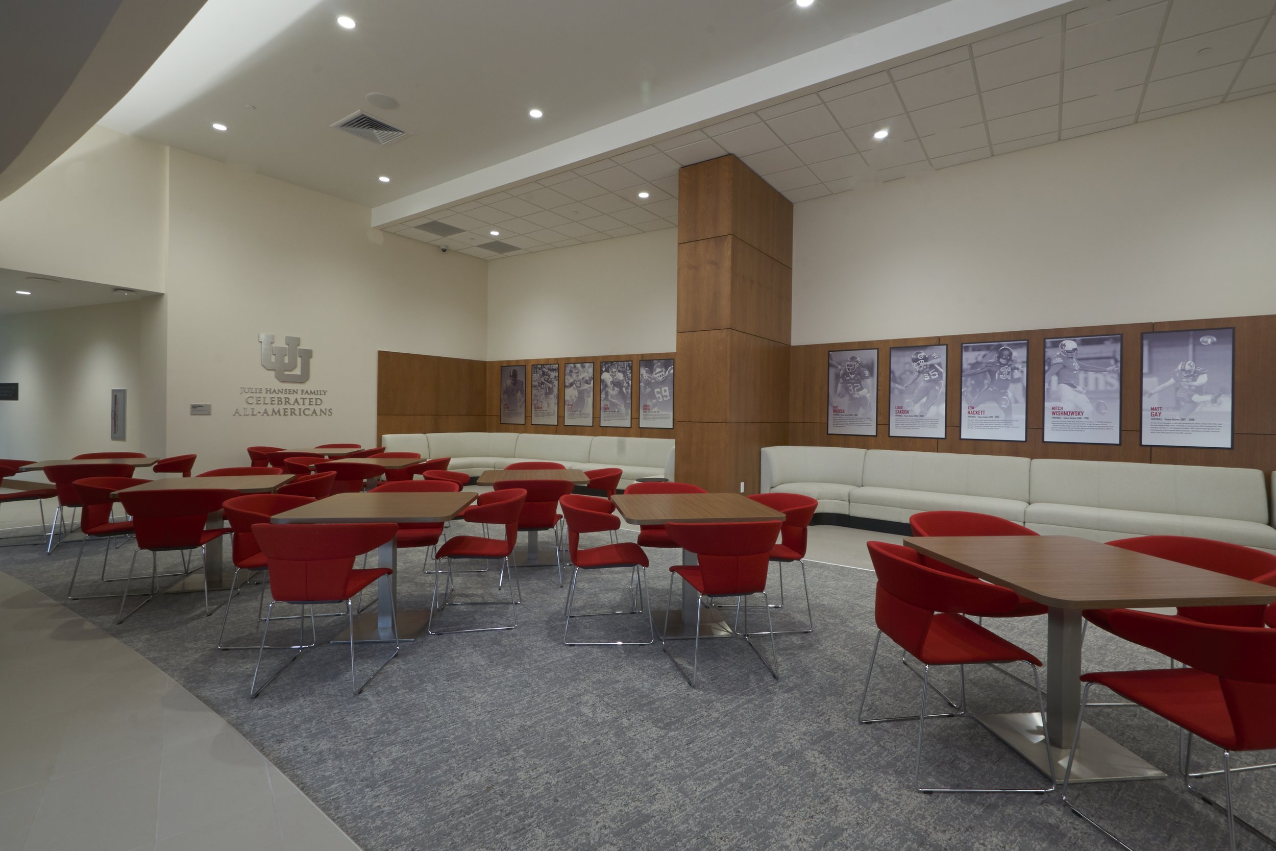 University of Indiana football lounge with red chairs, wooden tables, white curved seating, framed football player posters on the wall, and the IU logo with the text celebrating all-Americans.