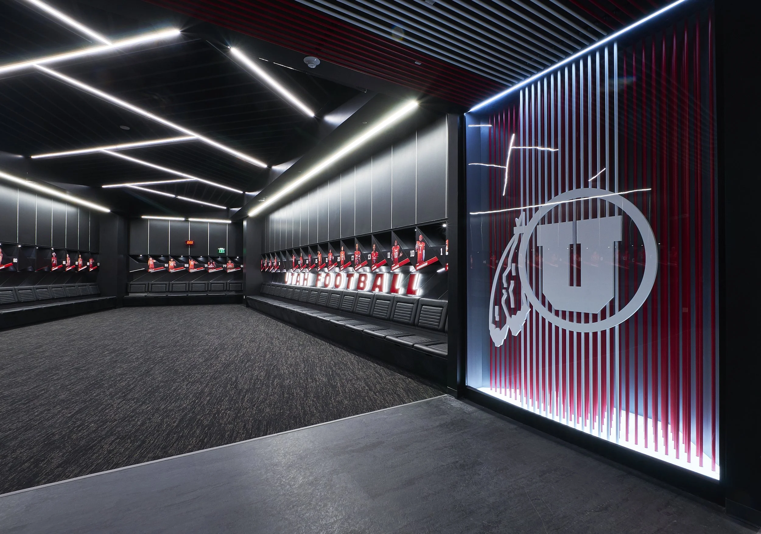Empty locker room with black benches and wall-mounted lockers, illuminated by modern linear ceiling lights, with a large Utah Football logo on a wall with vertical red stripes.