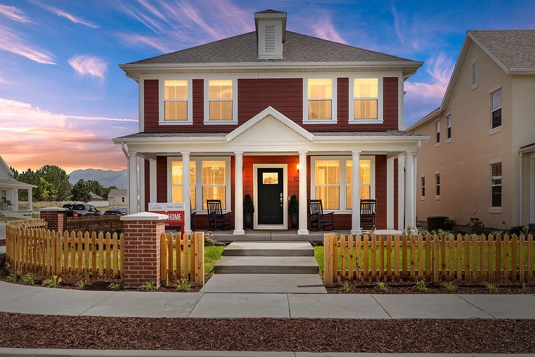 Two-story red house with white trim and porch at sunset, featuring a picket fence, green lawn, and rocking chairs.