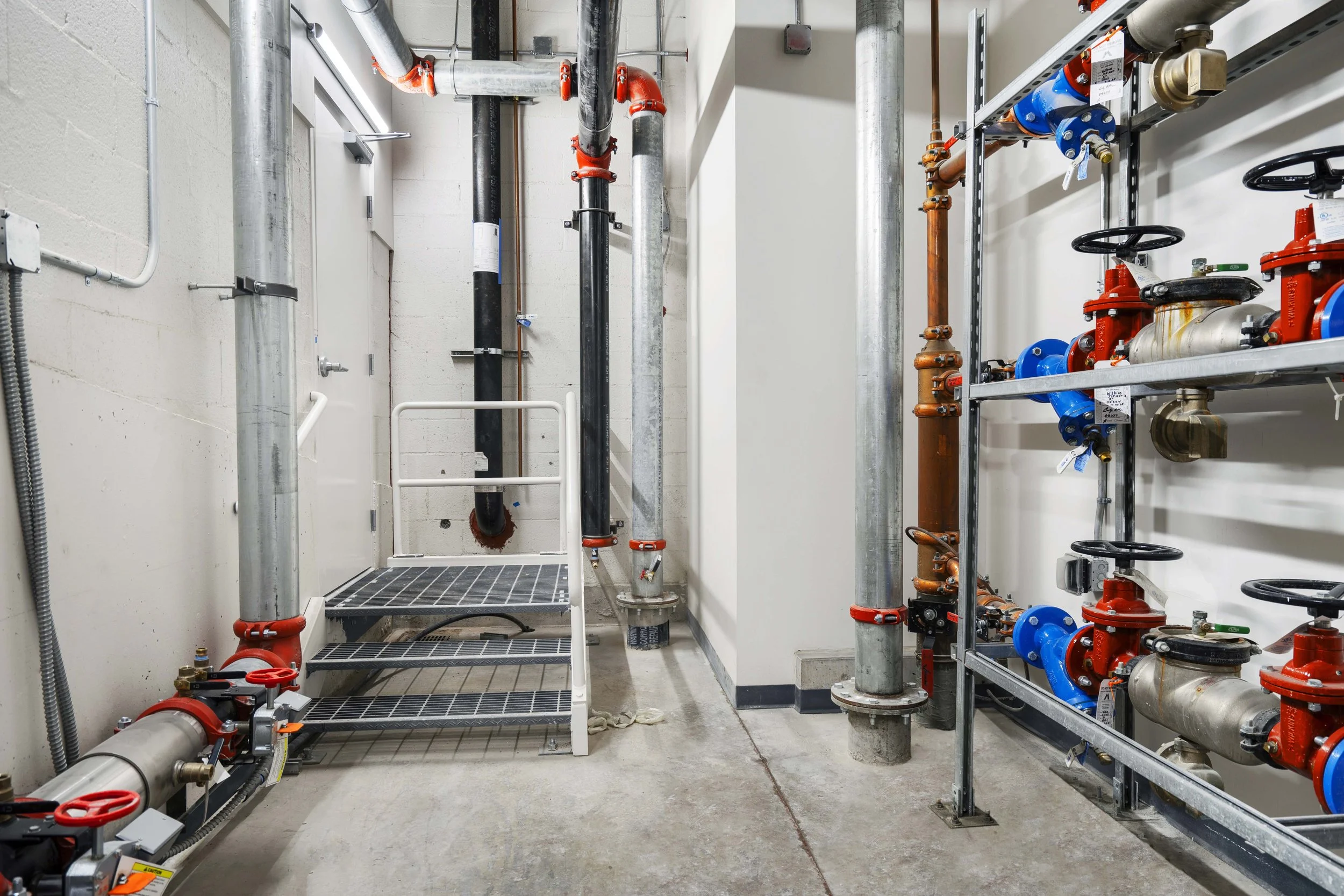 A utility room with pipes, valves, and metal shelving, including a small metal staircase.