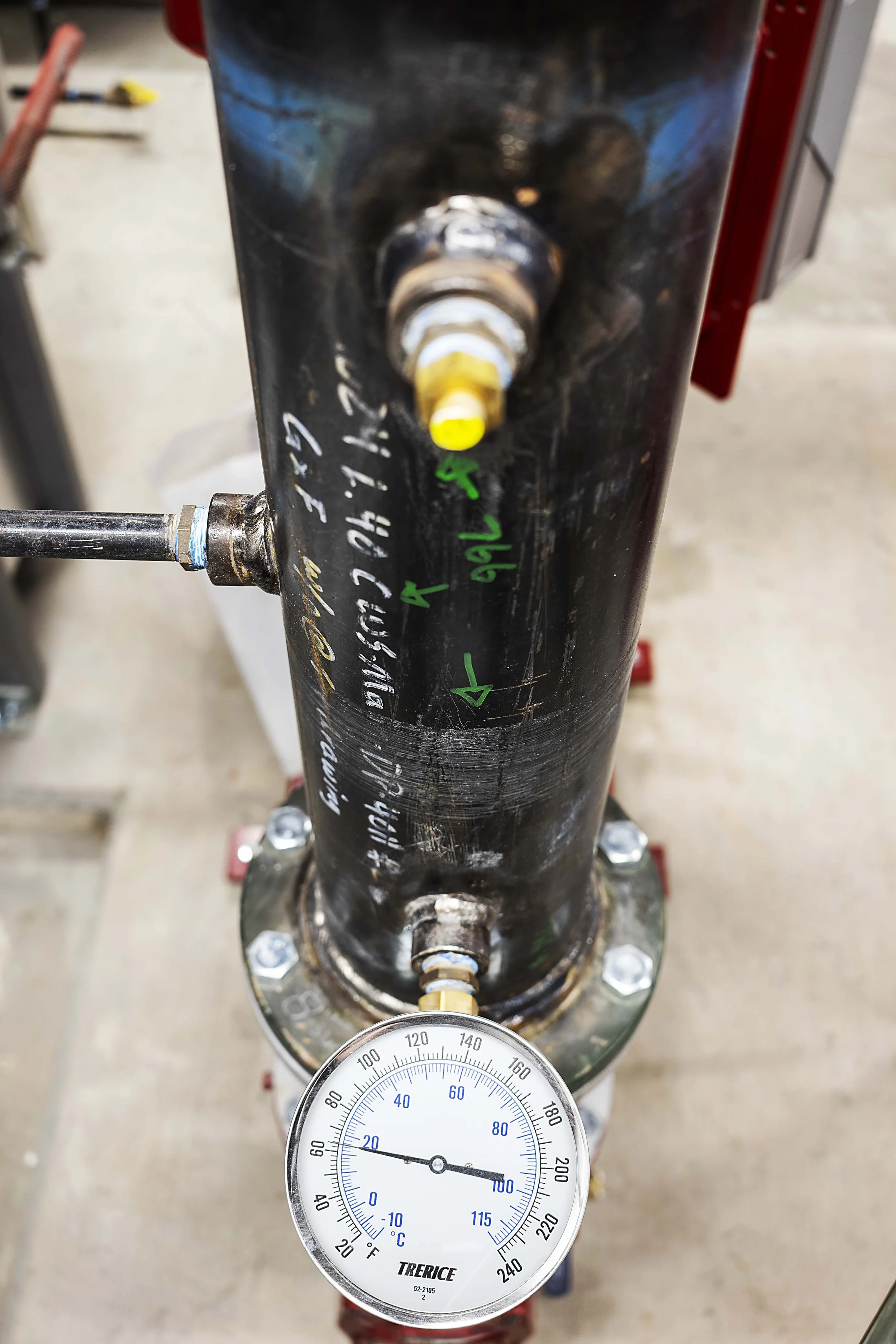 View looking down at a black pipe with handwritten notes and a pressure gauge attached, in a workshop or industrial setting.