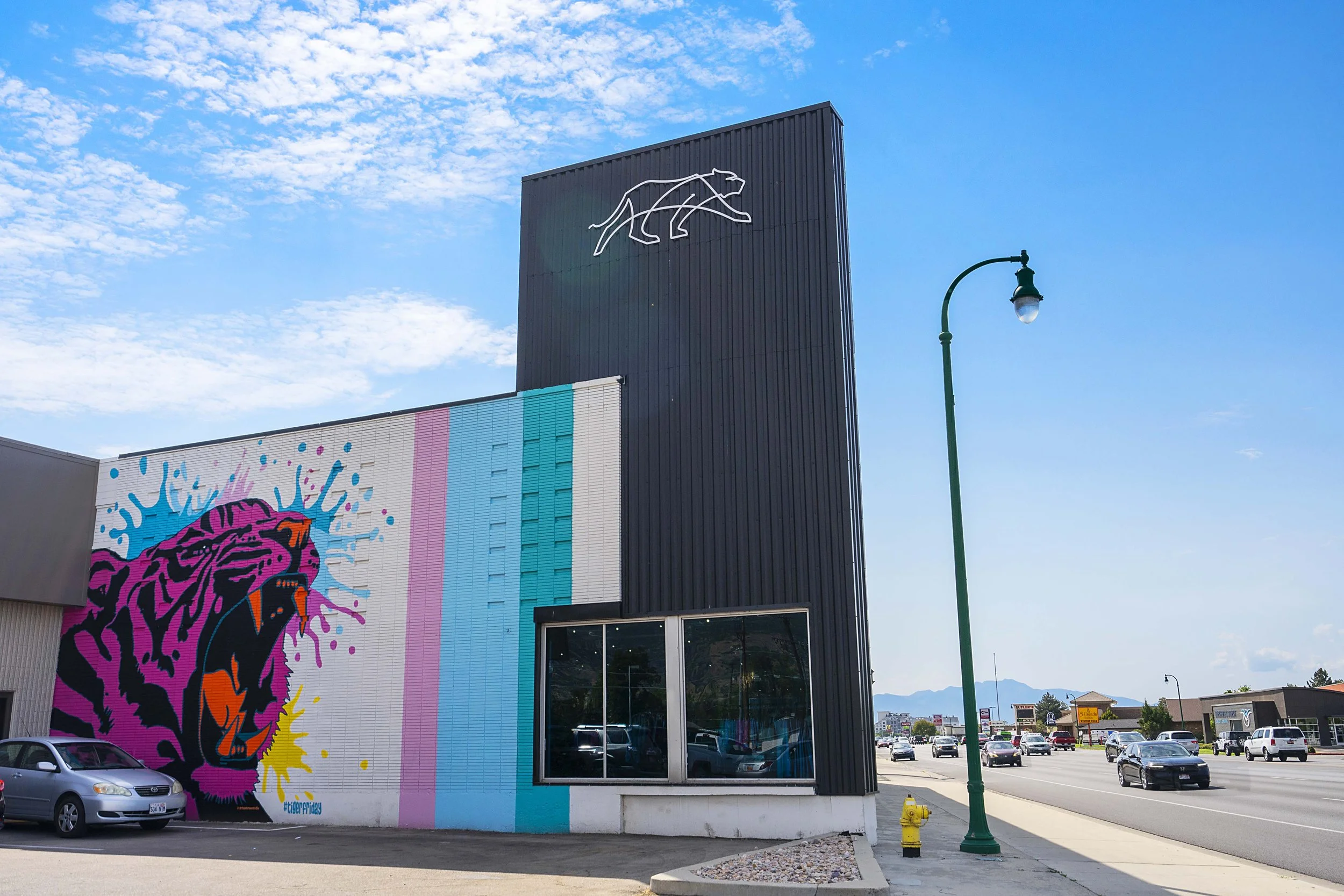 A building with a large colorful mural of a roaring pink and black tiger with splashes of blue, yellow, and pink paint, and a white outline of a lion on a black tower. The scene includes a street with cars, a green streetlamp, and a blue sky with sca