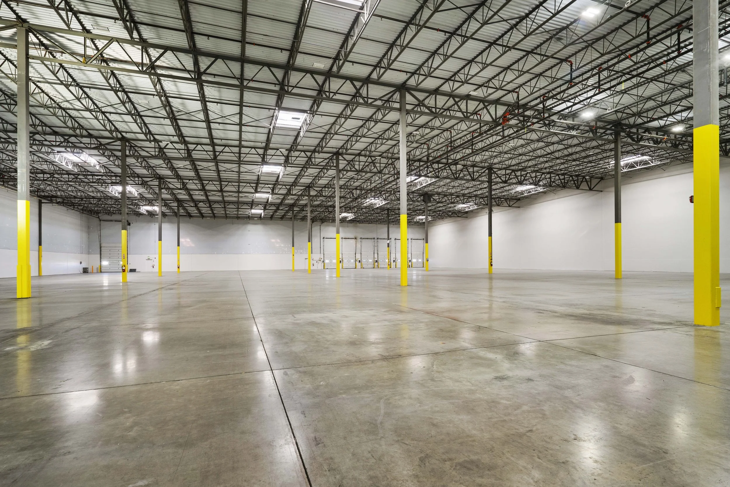 Empty industrial warehouse with yellow support columns, a high ceiling with exposed metal framework, and a polished concrete floor.