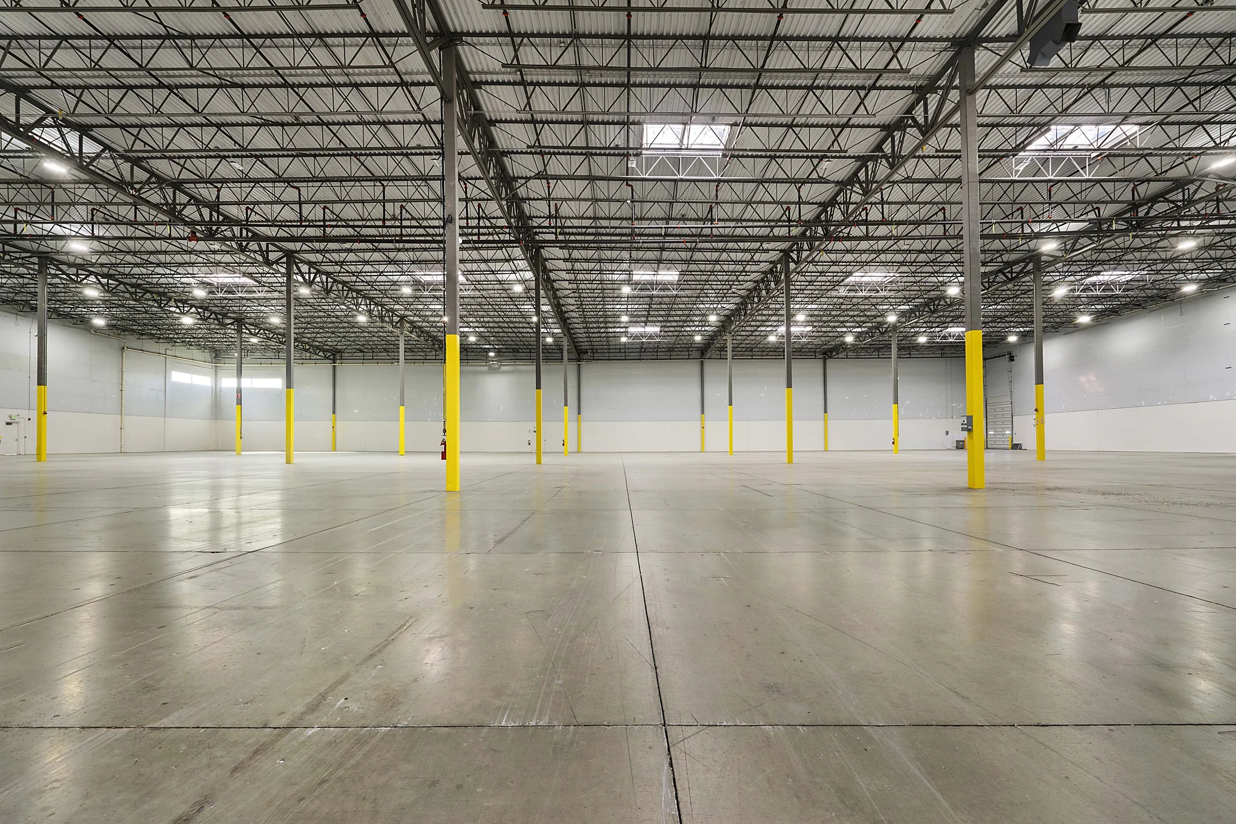 Empty indoor warehouse with concrete floor, yellow support columns, and a ceiling with exposed metal trusses and skylights.