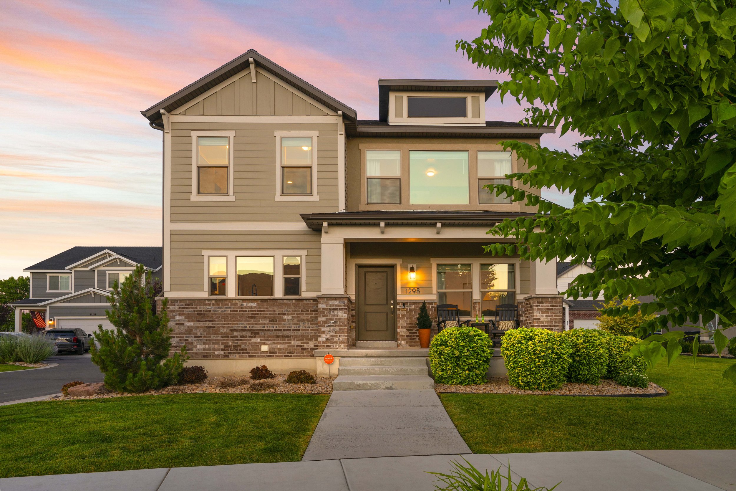 A modern two-story house with gray siding, brick foundation, and a front porch with outdoor seating, illuminated by a porch light, during sunset with colorful sky and green trees.