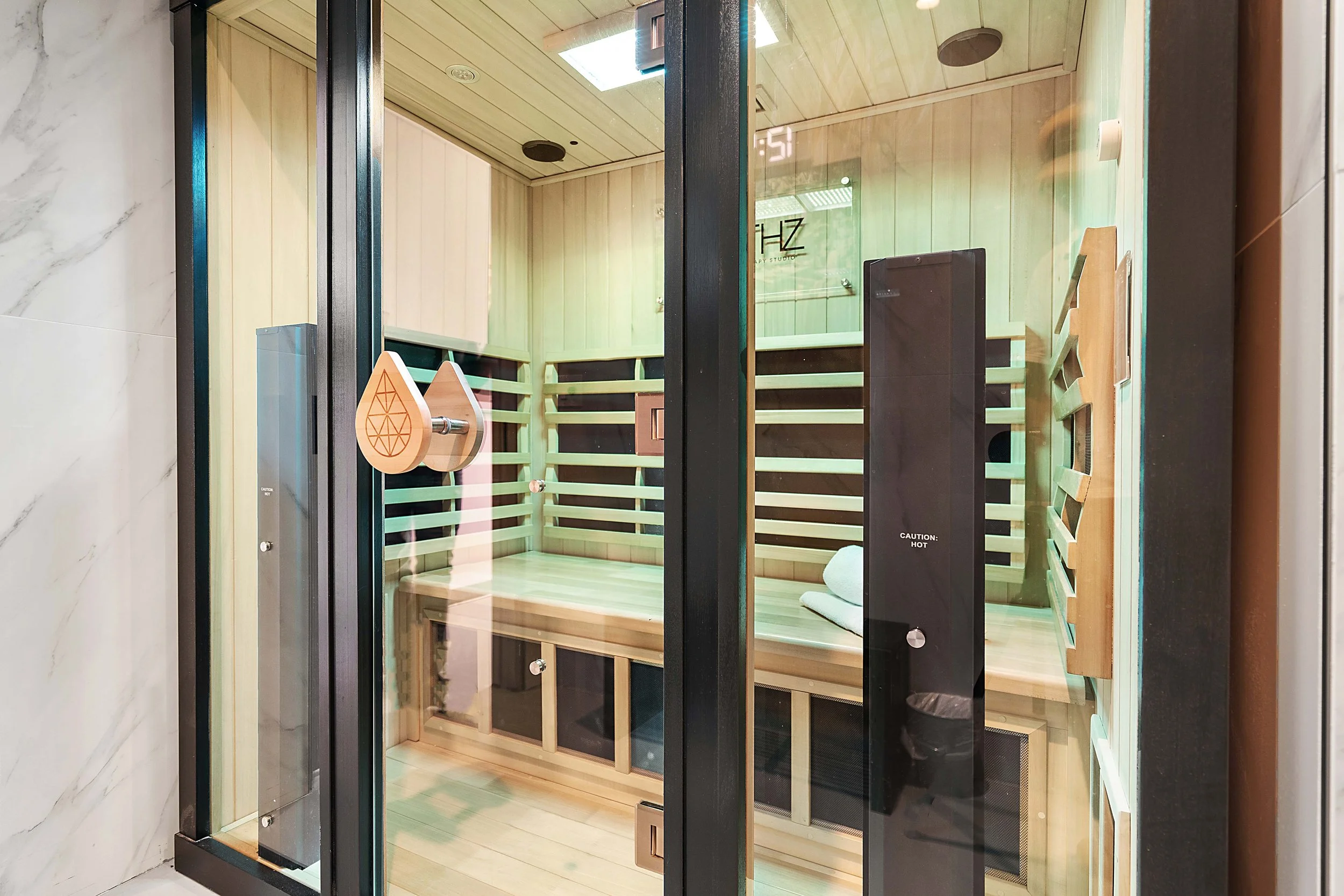 An infrared sauna chamber with a wooden interior, black frame, and glass walls. There are two teardrop-shaped wooden ornaments hanging on the door handle and a towel with a small cushion inside.