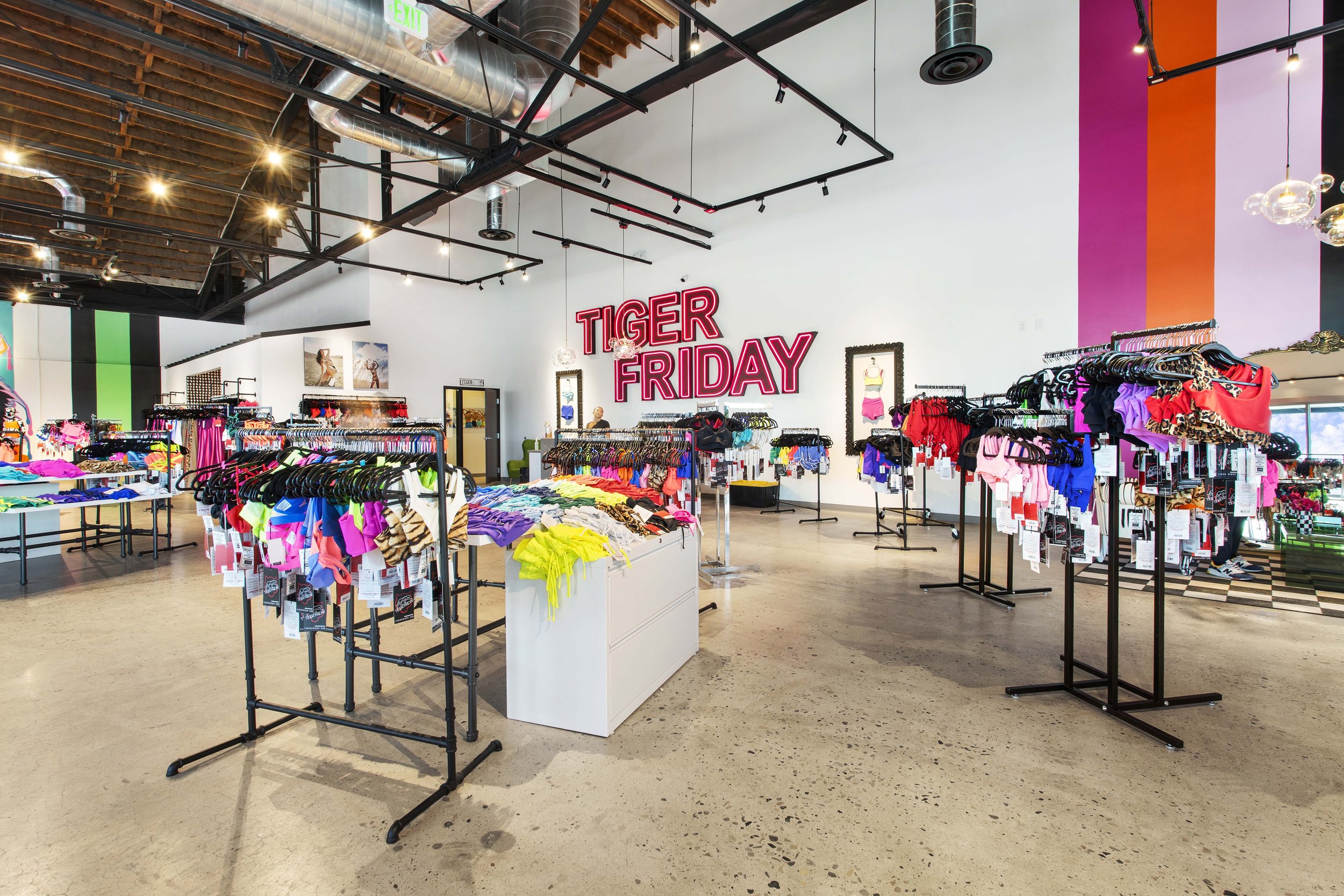 A clothing store featuring racks of brightly colored swimwear and apparel, with a neon sign that reads 'TIGER FRIDAY' on the white wall behind. The store has a modern industrial design with exposed ceiling pipes, track lighting, and a polished concre