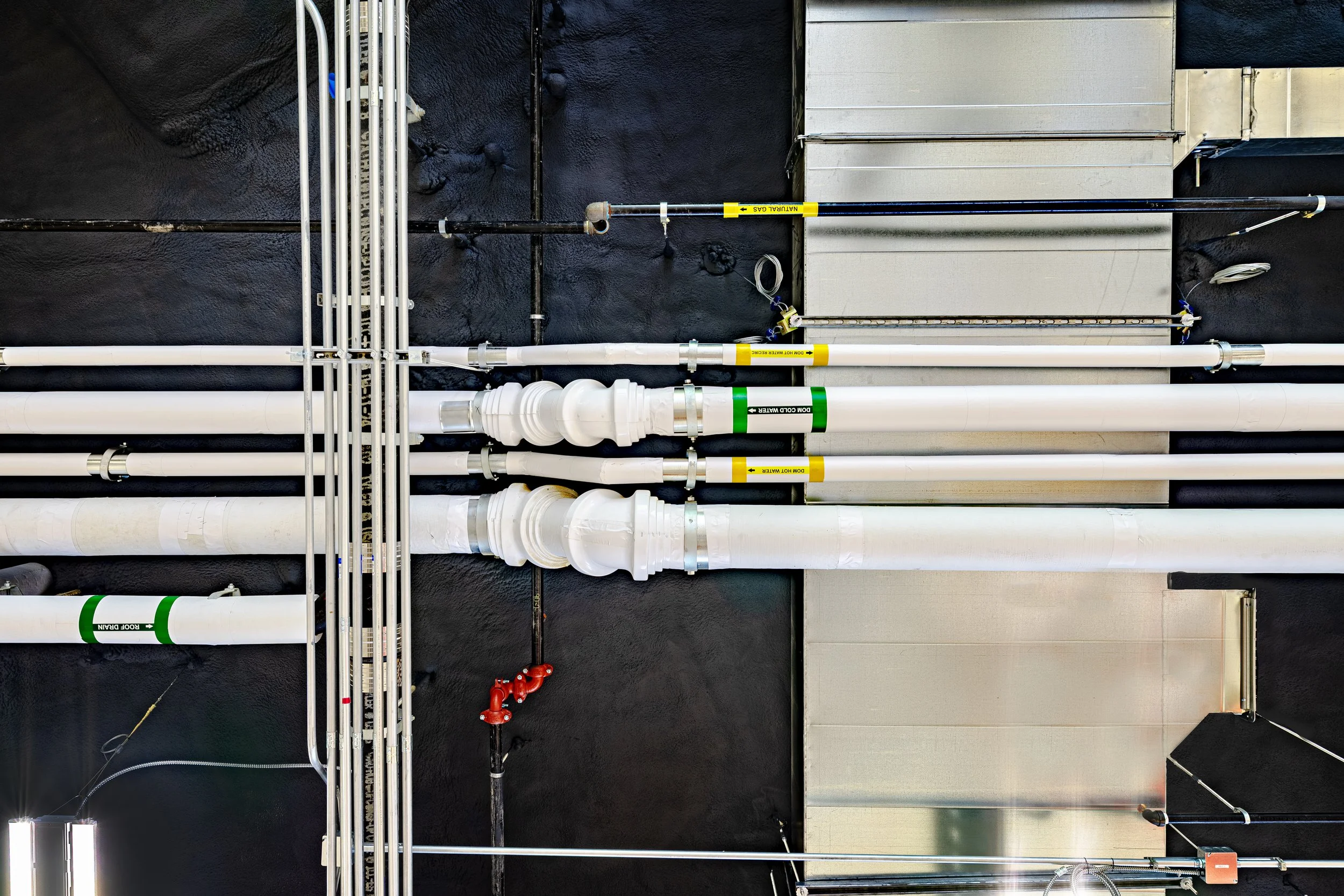 Industrial piping system with white pipes, black pipes, labels, and valves on a dark wall.
