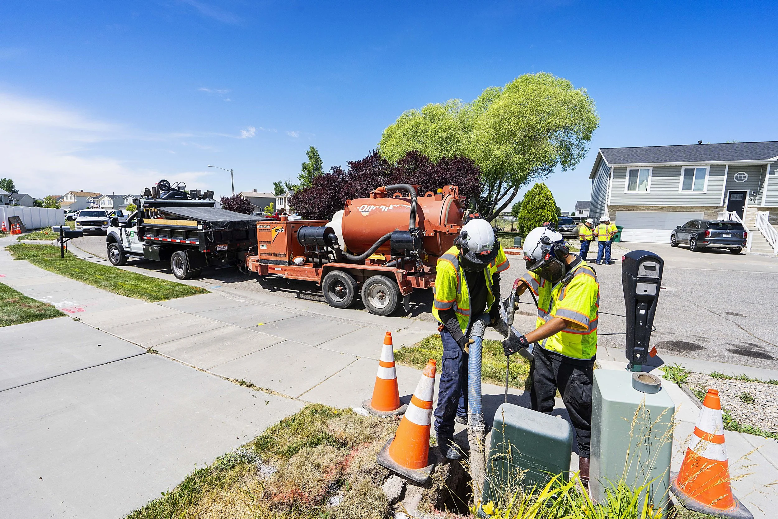 Two workers in yellow high-visibility shirts and helmets are working on a utility box with tools on a sidewalk, with traffic cones around them. In the background, there are street workers, houses, and a large maintenance truck with a vacuum system on