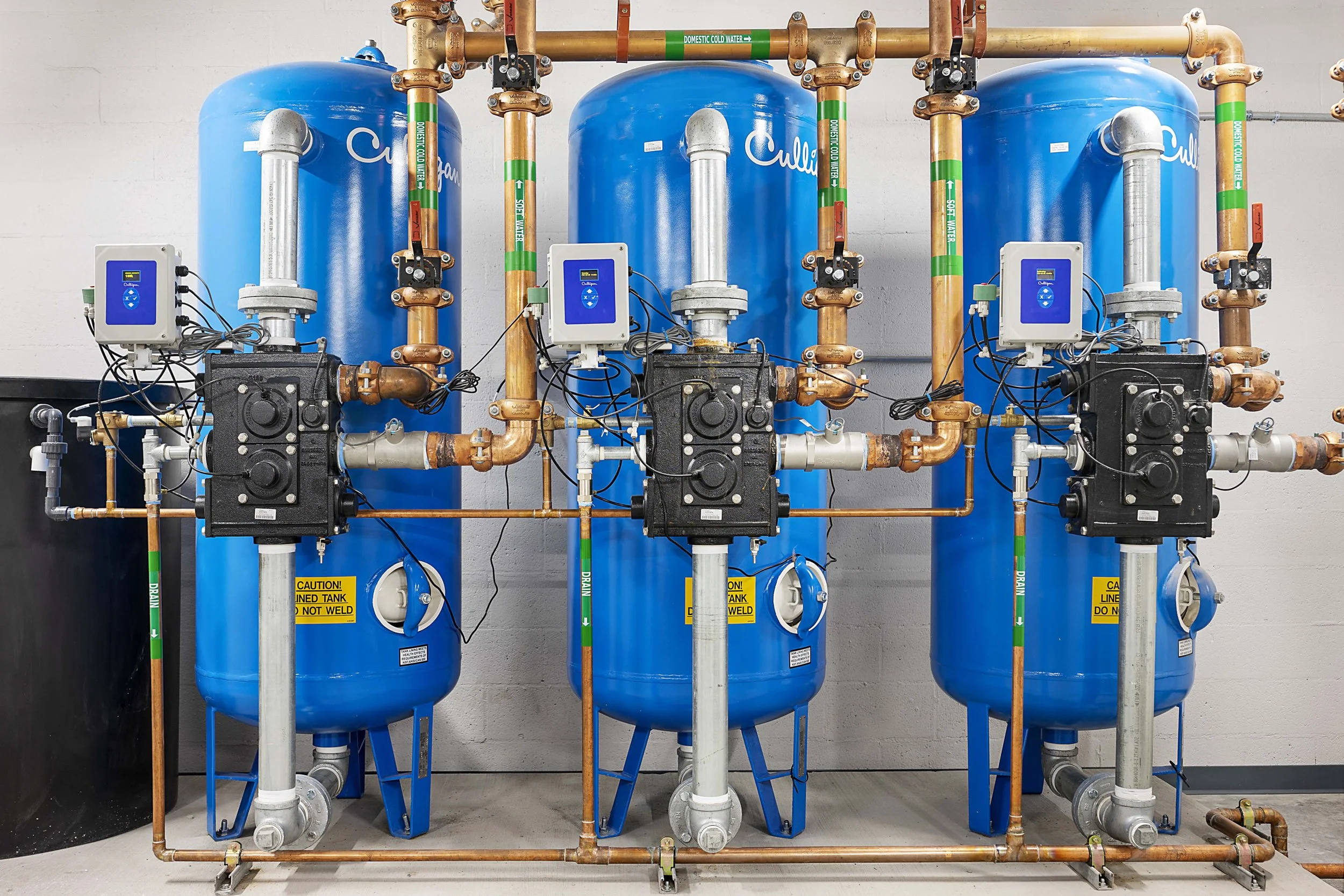 Three large blue water filtration tanks connected by a network of copper pipes and black control boxes, with digital controls on top, mounted on a gray concrete wall in a utility room.