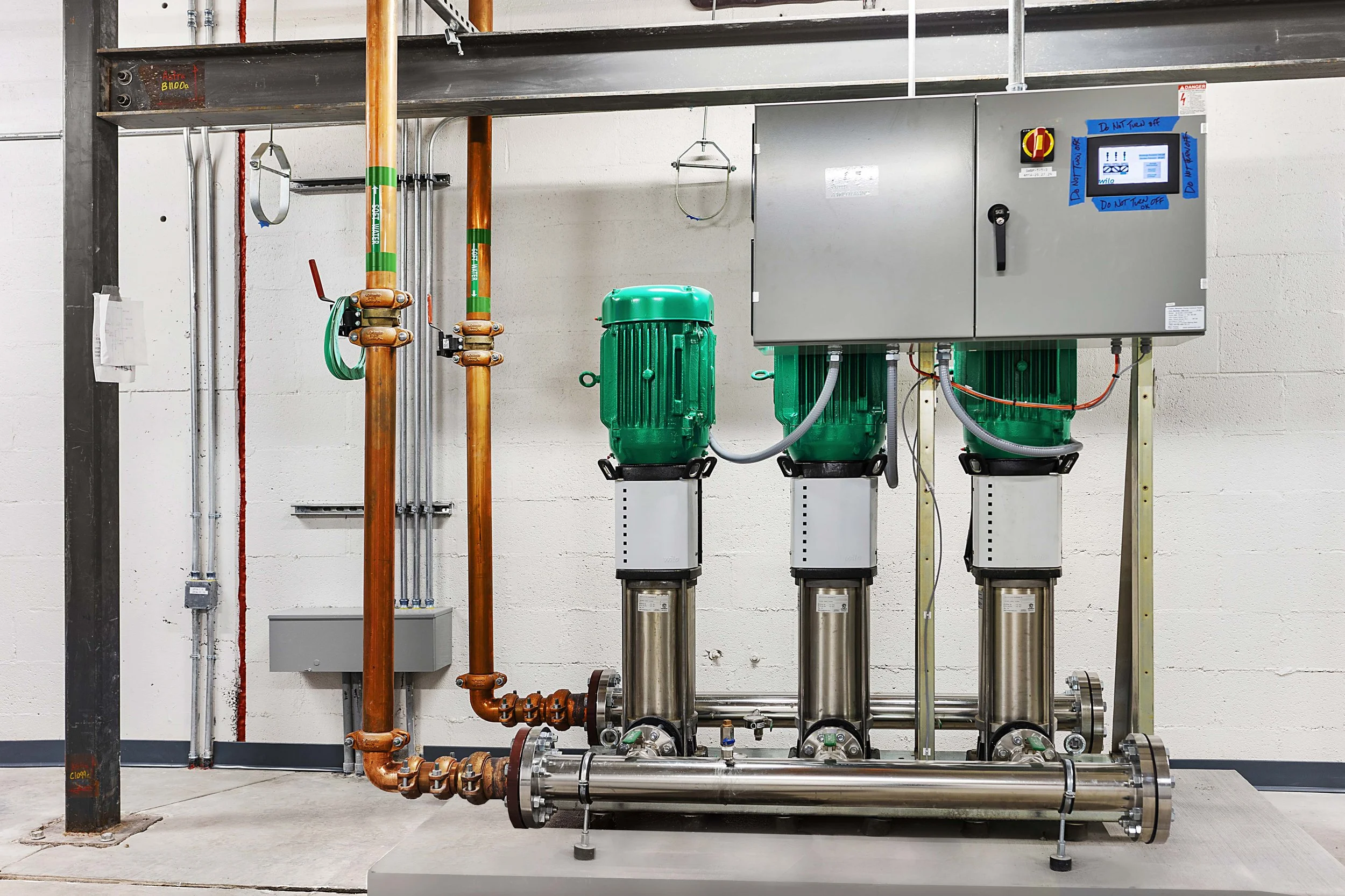 Industrial water pump system with green motors, pipes, and control panel mounted on a metal stand against a white brick wall.