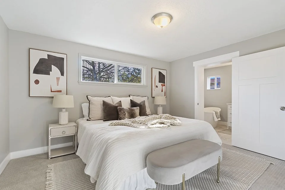 A cozy bedroom with a white bed, beige and brown pillows, and abstract artwork on the walls. There's a small bench at the foot of the bed and a view of a bathroom in the background.
