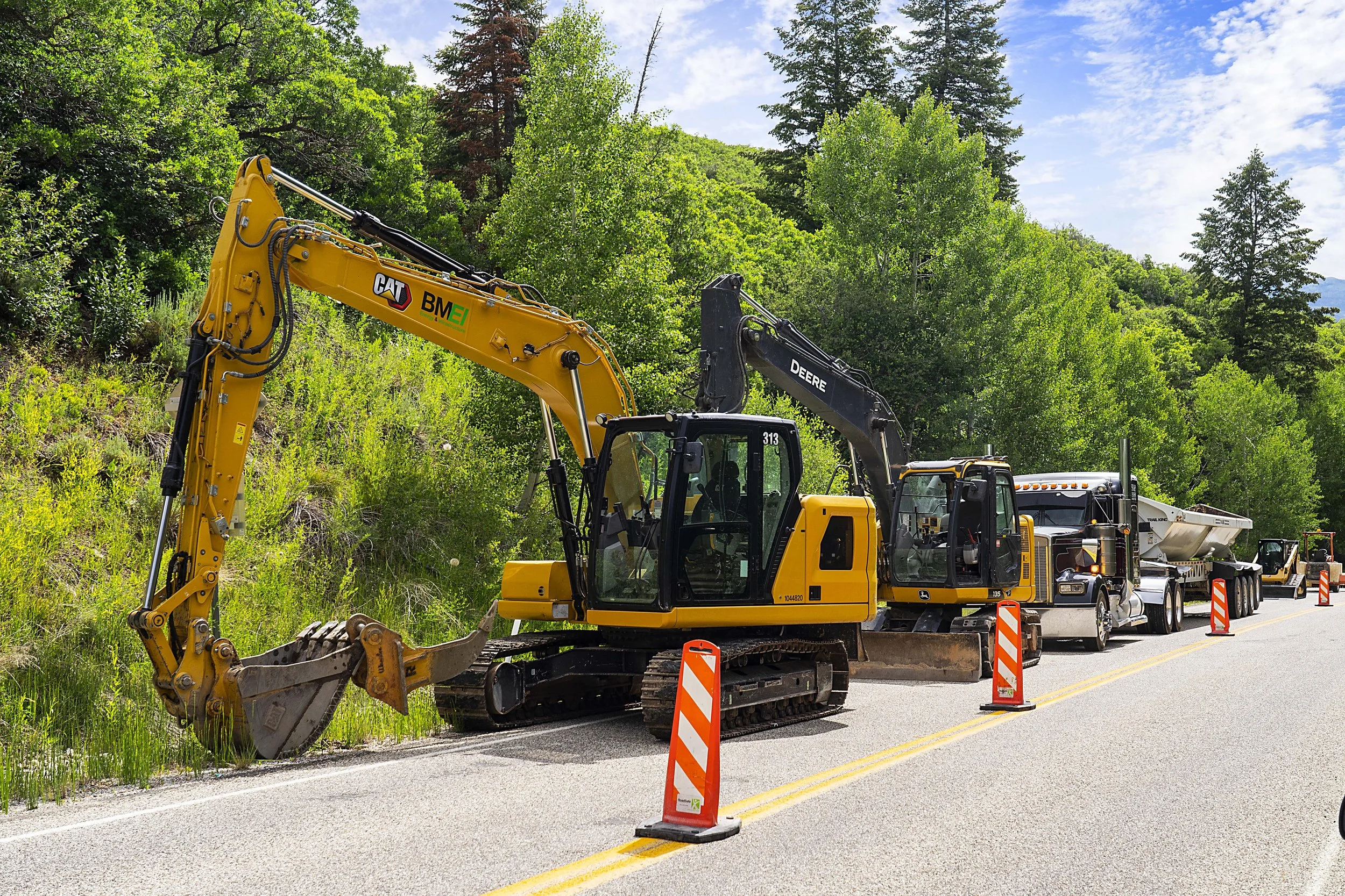 Construction vehicles working on the side of a rural road surrounded by green trees and forest, with cones and barriers blocking part of the road for the work.