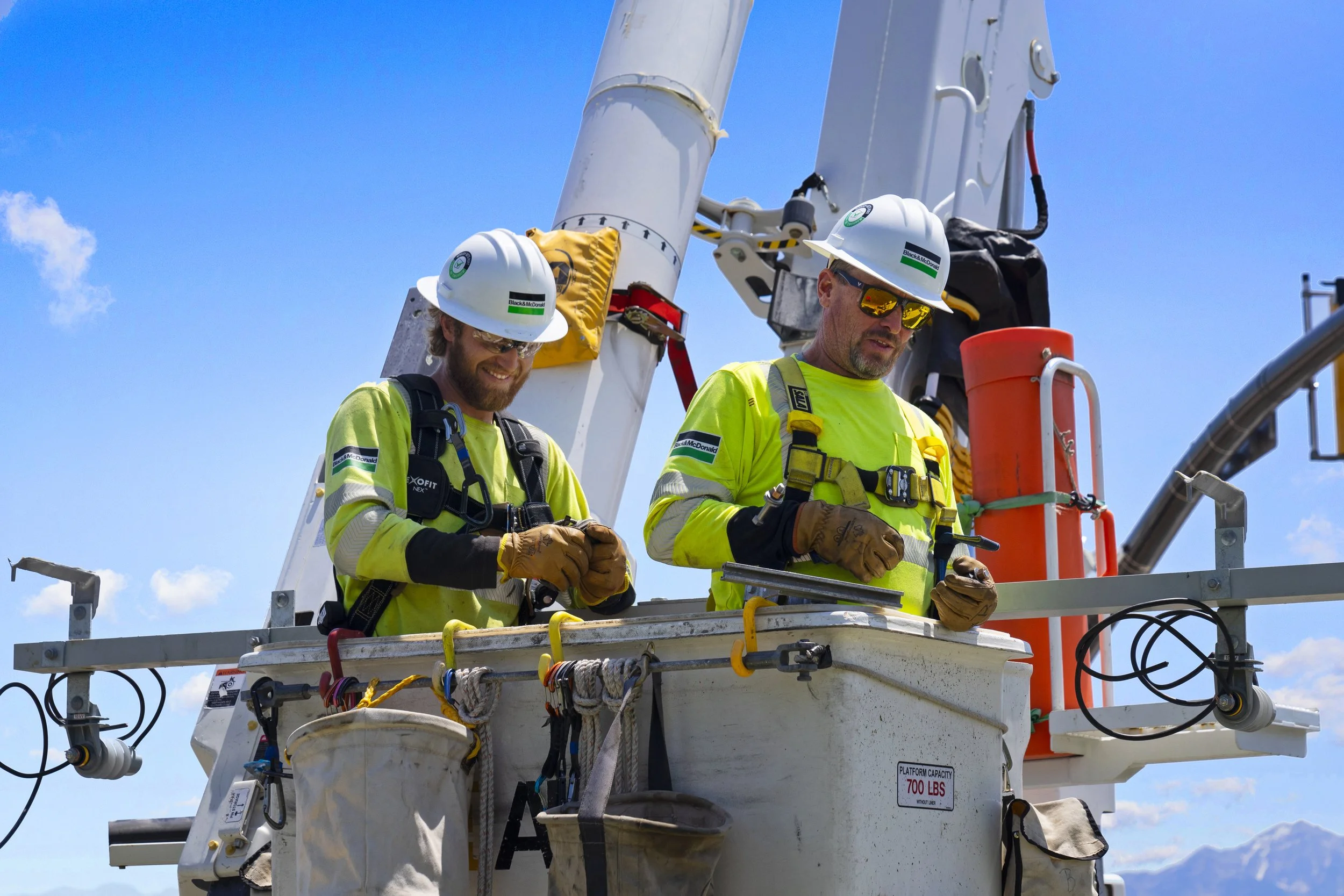 Two workers in safety gear and helmets standing in a lift basket working on electrical or construction equipment against a blue sky.