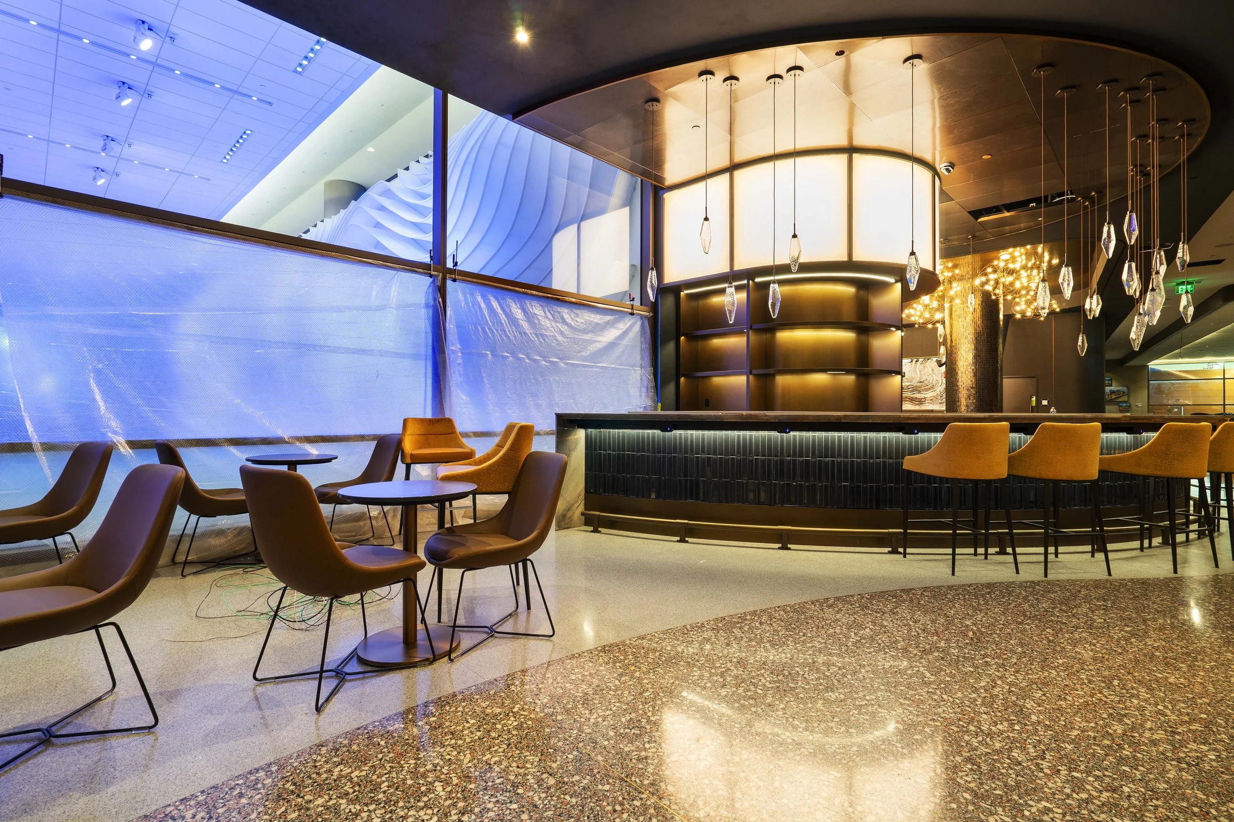 Interior of a modern bar with high bar stools, tables, and lounge chairs in a well-lit space featuring artistic lighting fixtures and large windows.