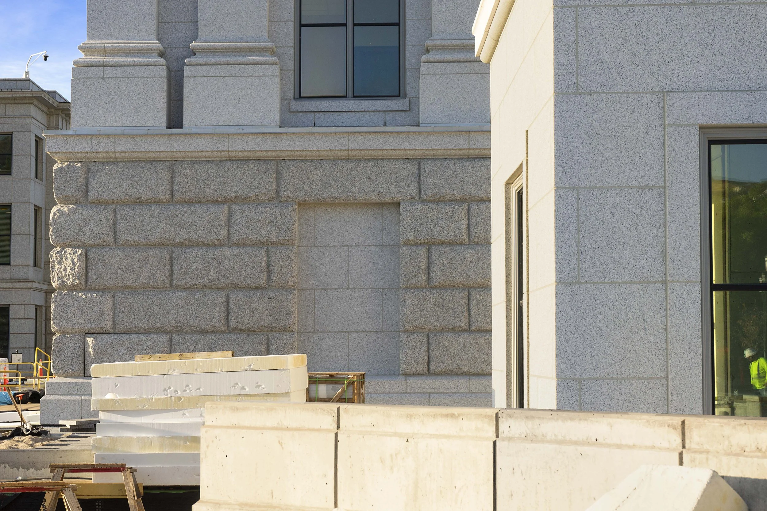 Construction site with a building featuring a stone exterior, large glass windows, and construction materials in the foreground.