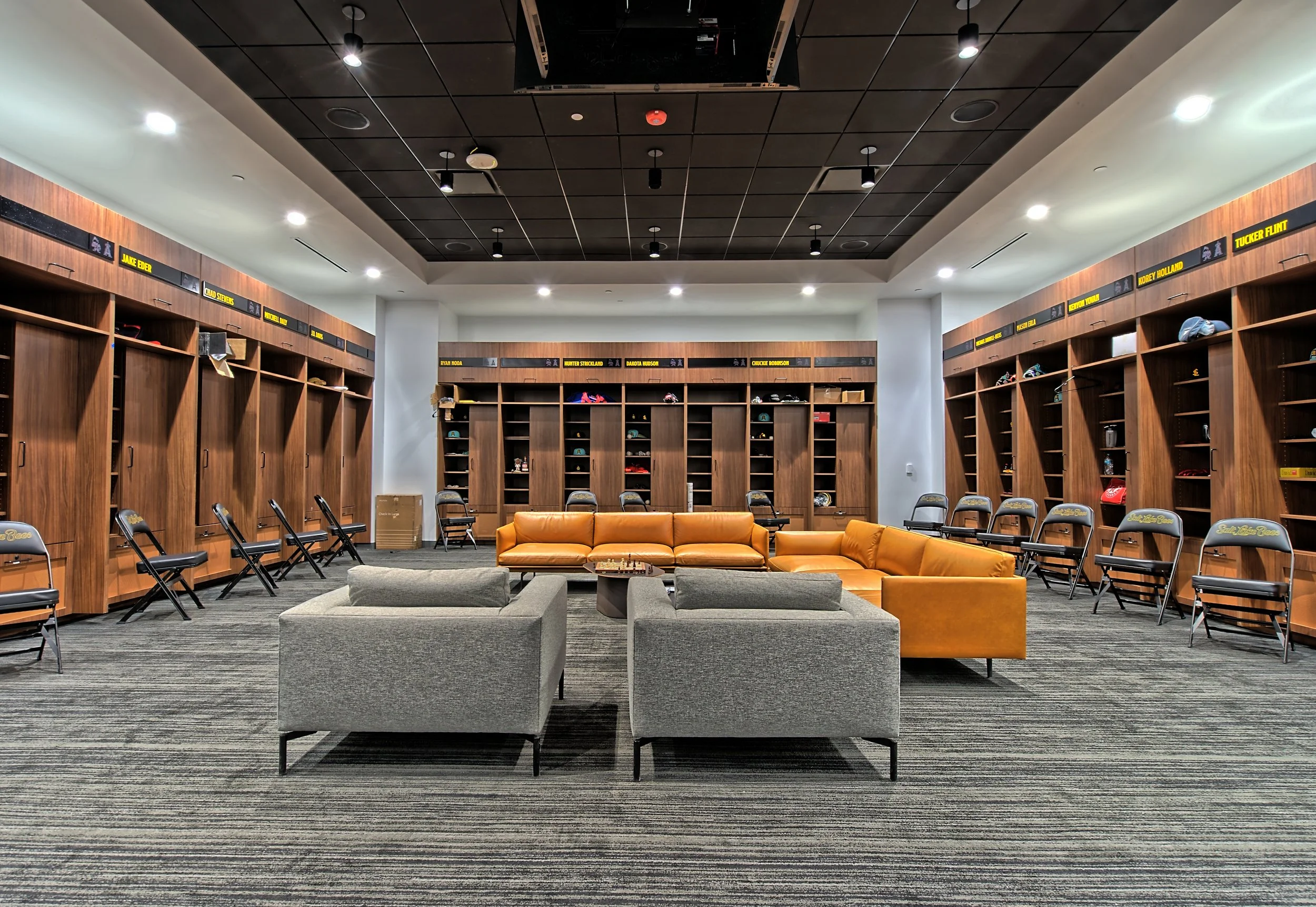 A spacious locker room with wooden lockers lining the walls, yellow and gray lounge chairs and sofas arranged in the center, and a striped carpeted floor. Overhead lighting illuminates the room.